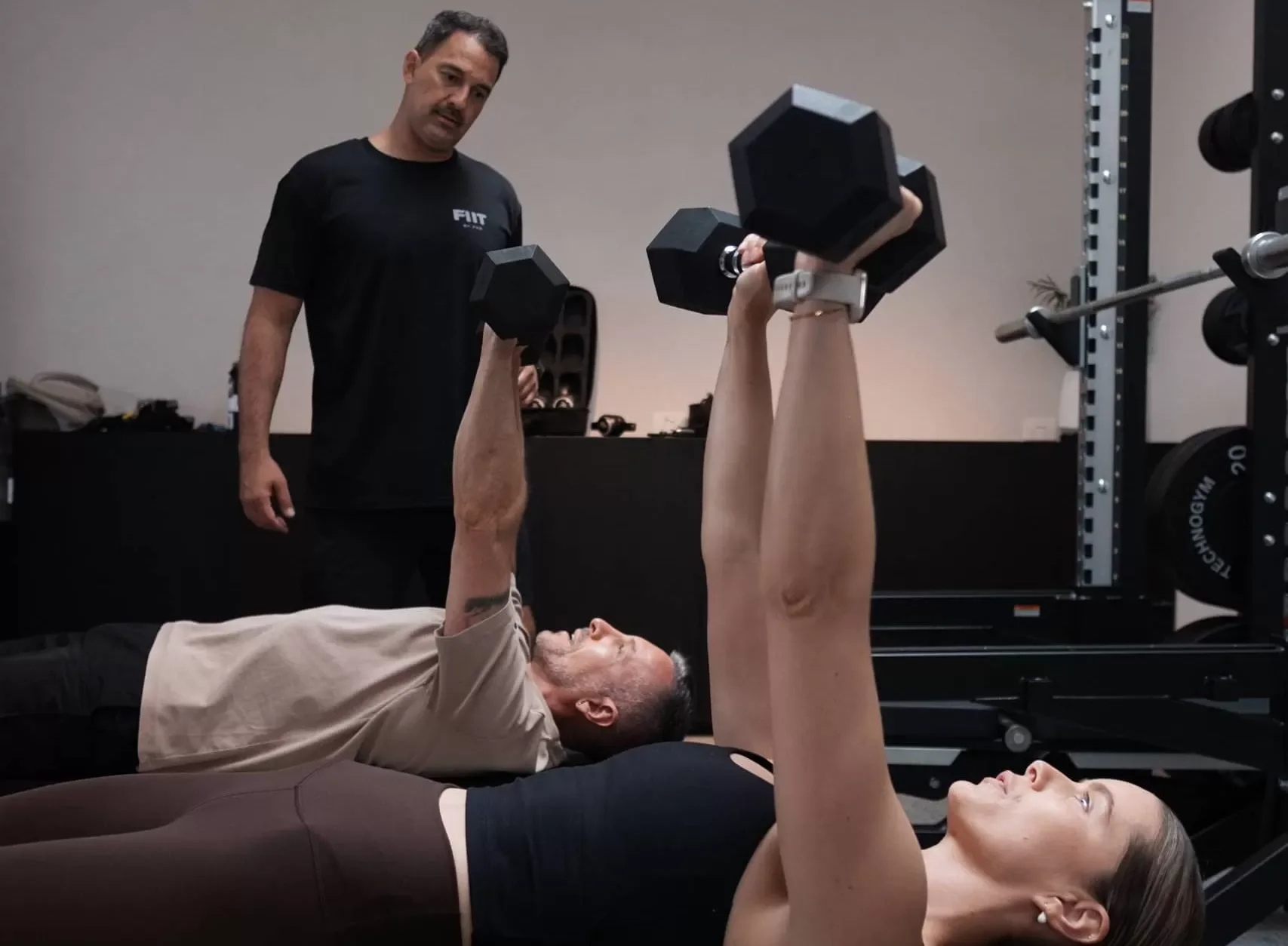 A woman lying on a bench performing a dumbbell chest press exercise with a man assisting her, while a trainer watches in the background in a gym.