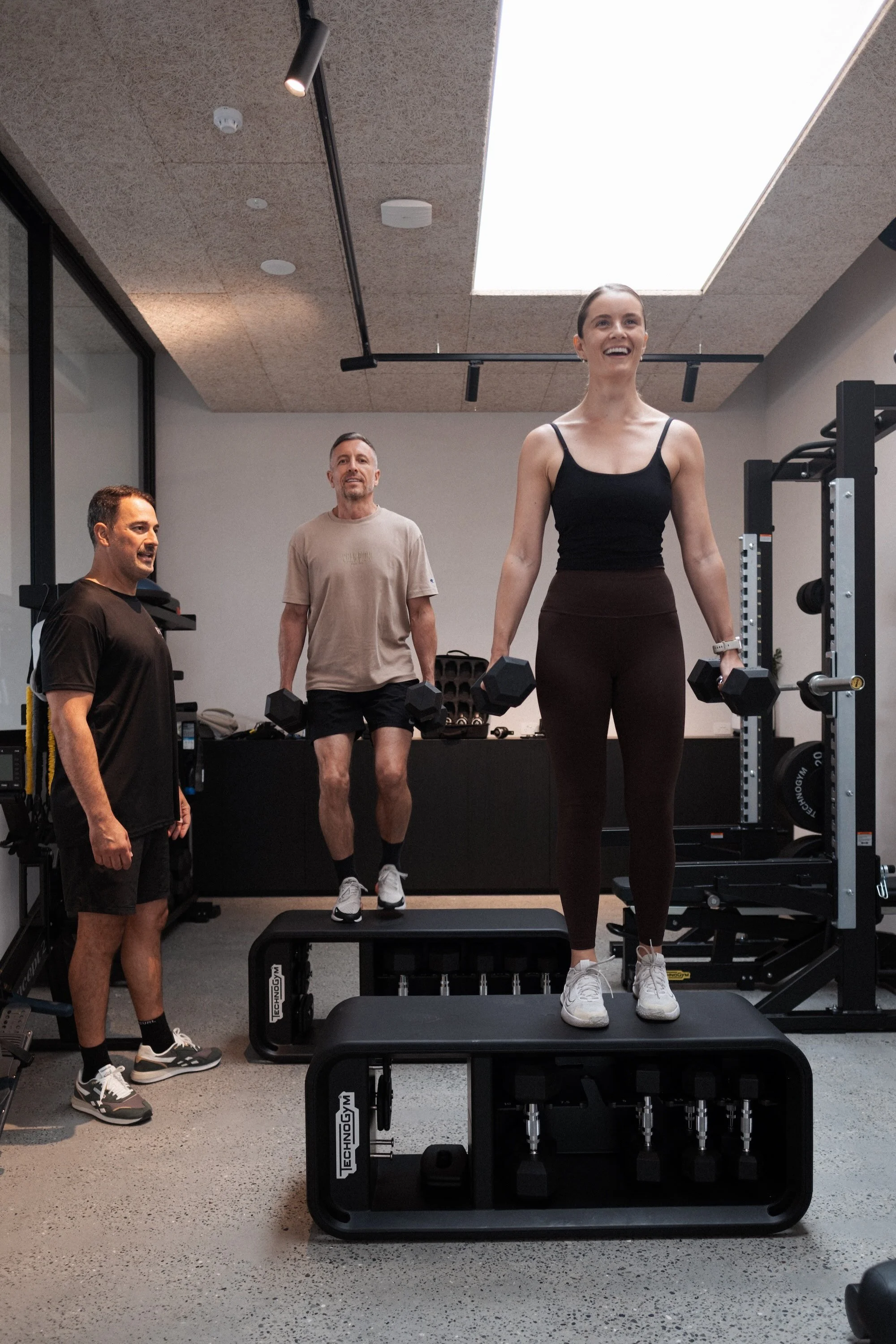 A woman holding dumbbells and smiling at a gym, with two men in the background conversing. Gym equipment and a skylight overhead.