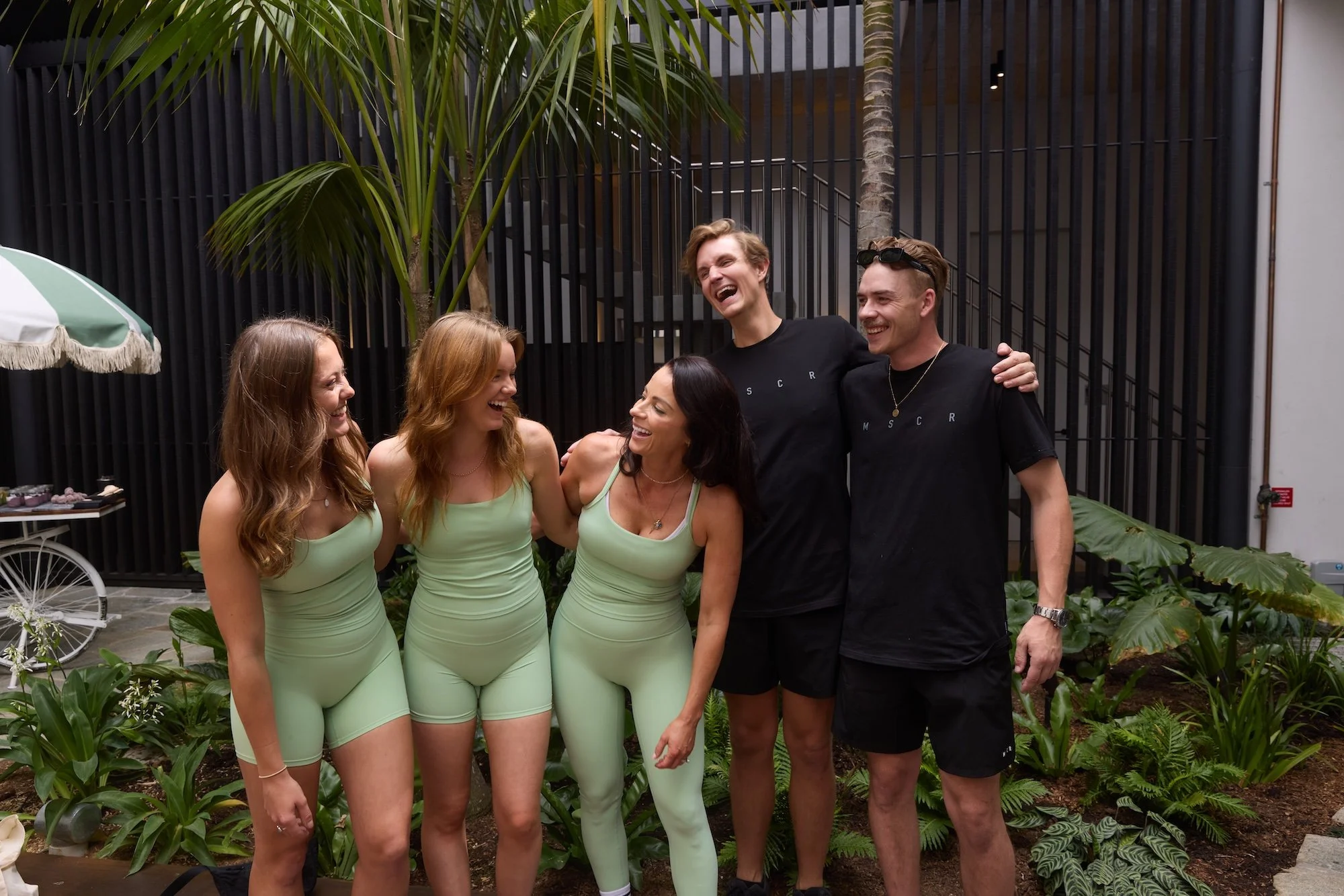 A group of six people, three women and three men, are standing together outdoors, smiling and laughing. The women are dressed in matching light green outfits, and the men are wearing black T-shirts with small white text. They appear to be enjoying a social gathering in a lush, green garden setting with various plants and tropical foliage.