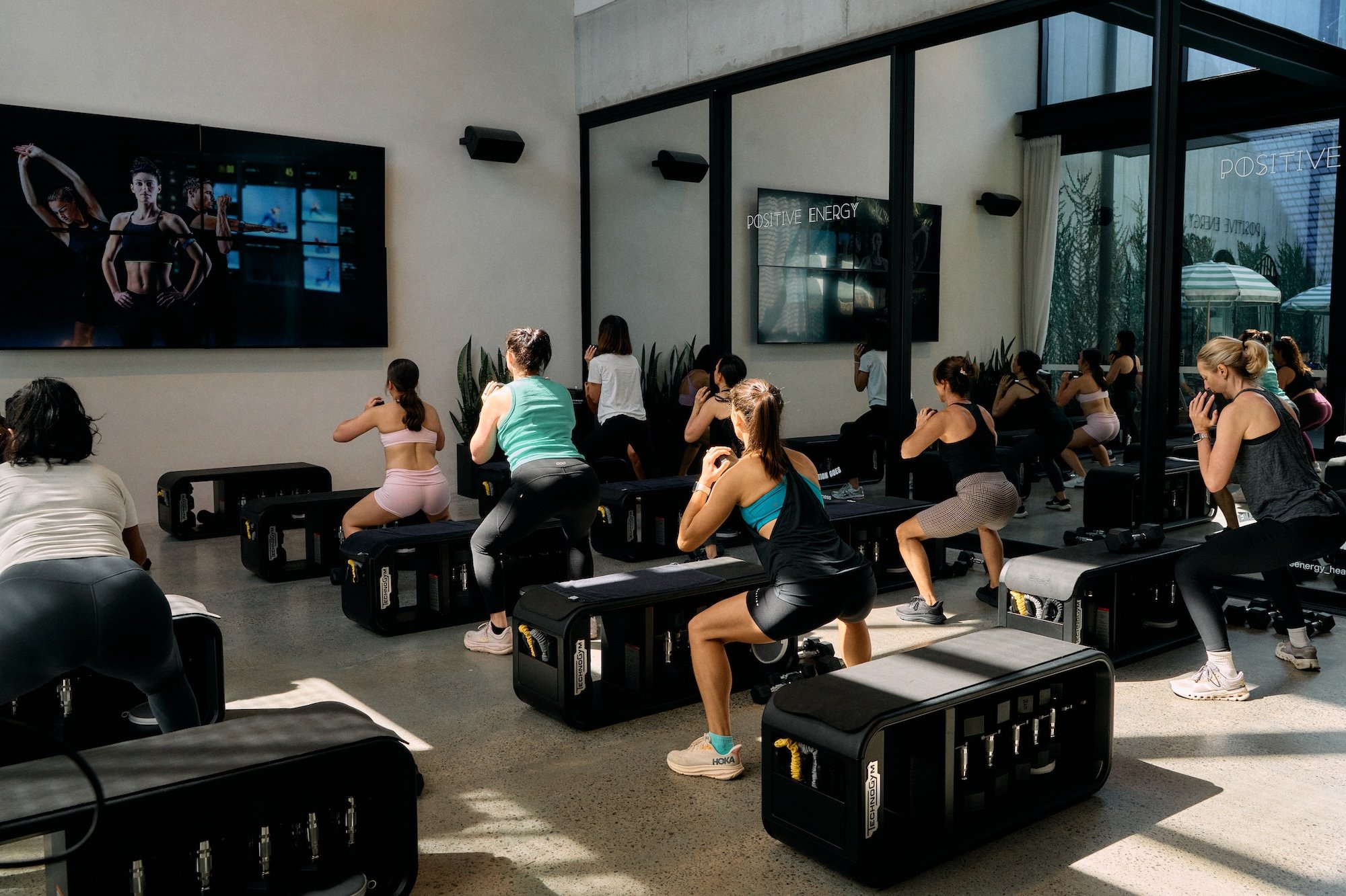 People participating in a group fitness class in a modern gym, performing squats with black workout benches and a large screen displaying fitness imagery in the background.