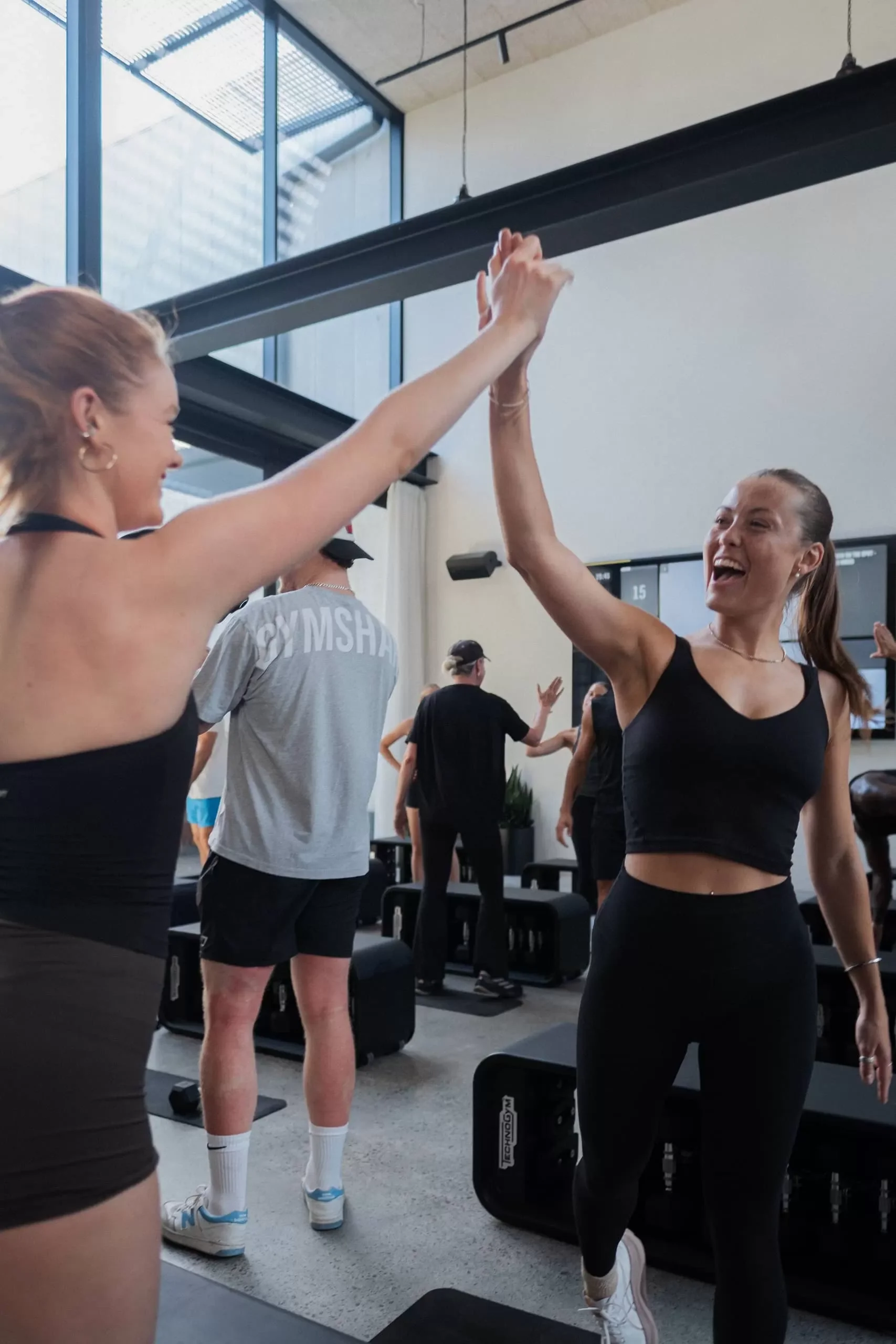 Women in workout clothes high-five each other in a fitness studio with large windows.