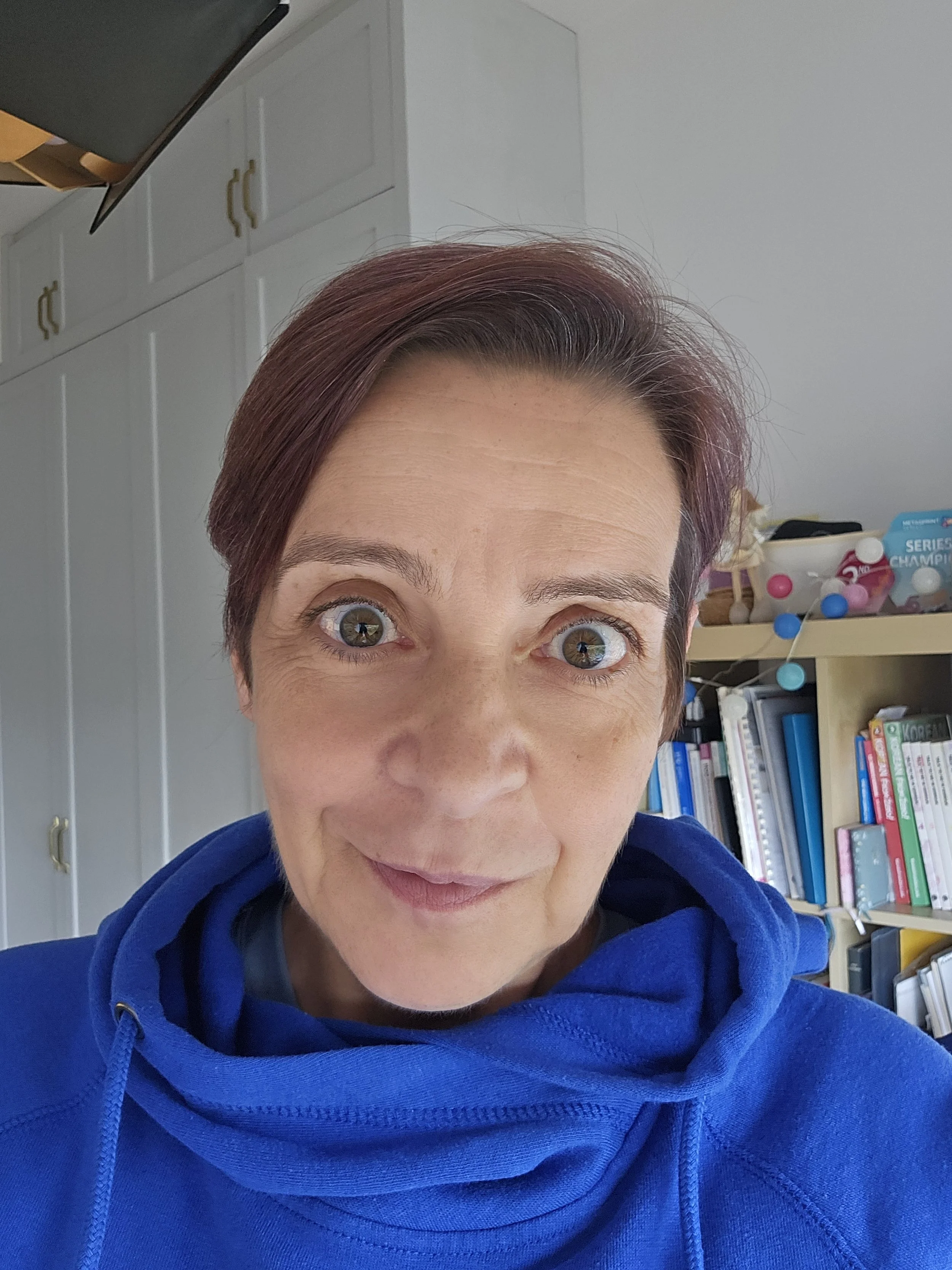 Close-up of a woman with short reddish-brown hair and blue eyes smiling, wearing a blue hoodie, in a room with white cabinets and bookshelves in the background.