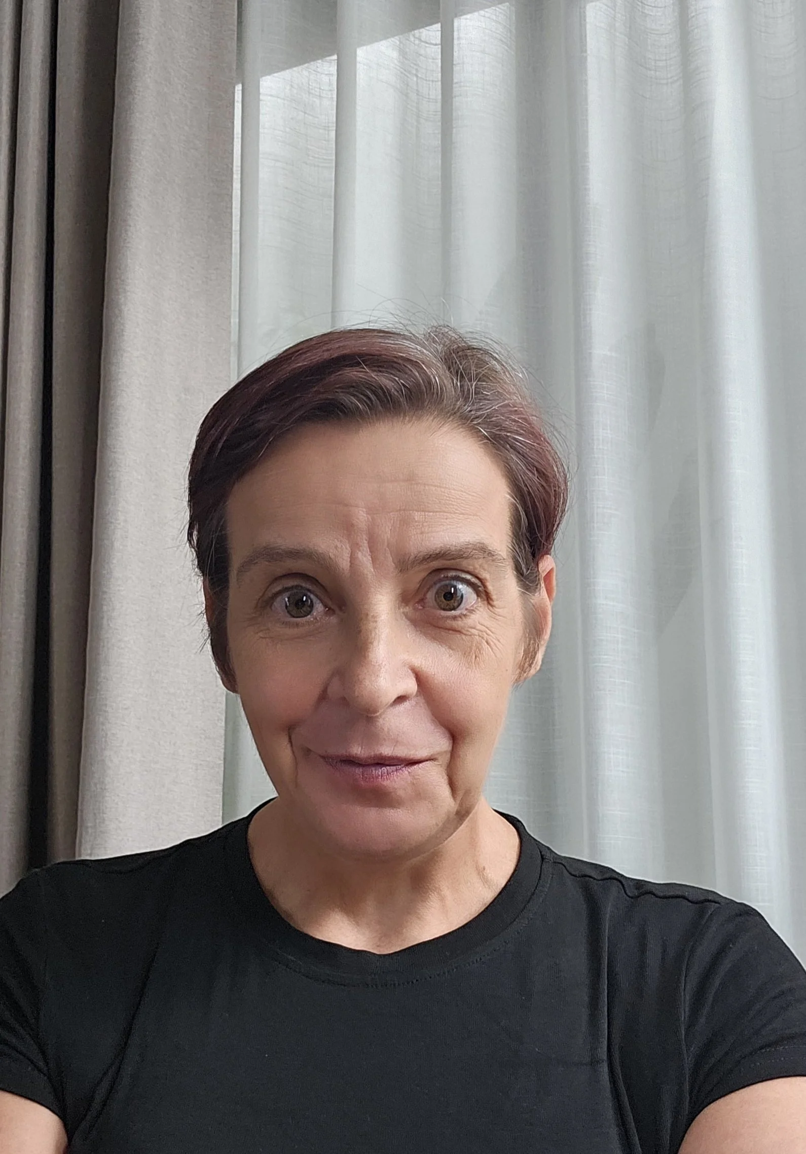 A woman with short, dark hair and bright eyes smiling, wearing a black shirt, standing in front of light-colored curtains.