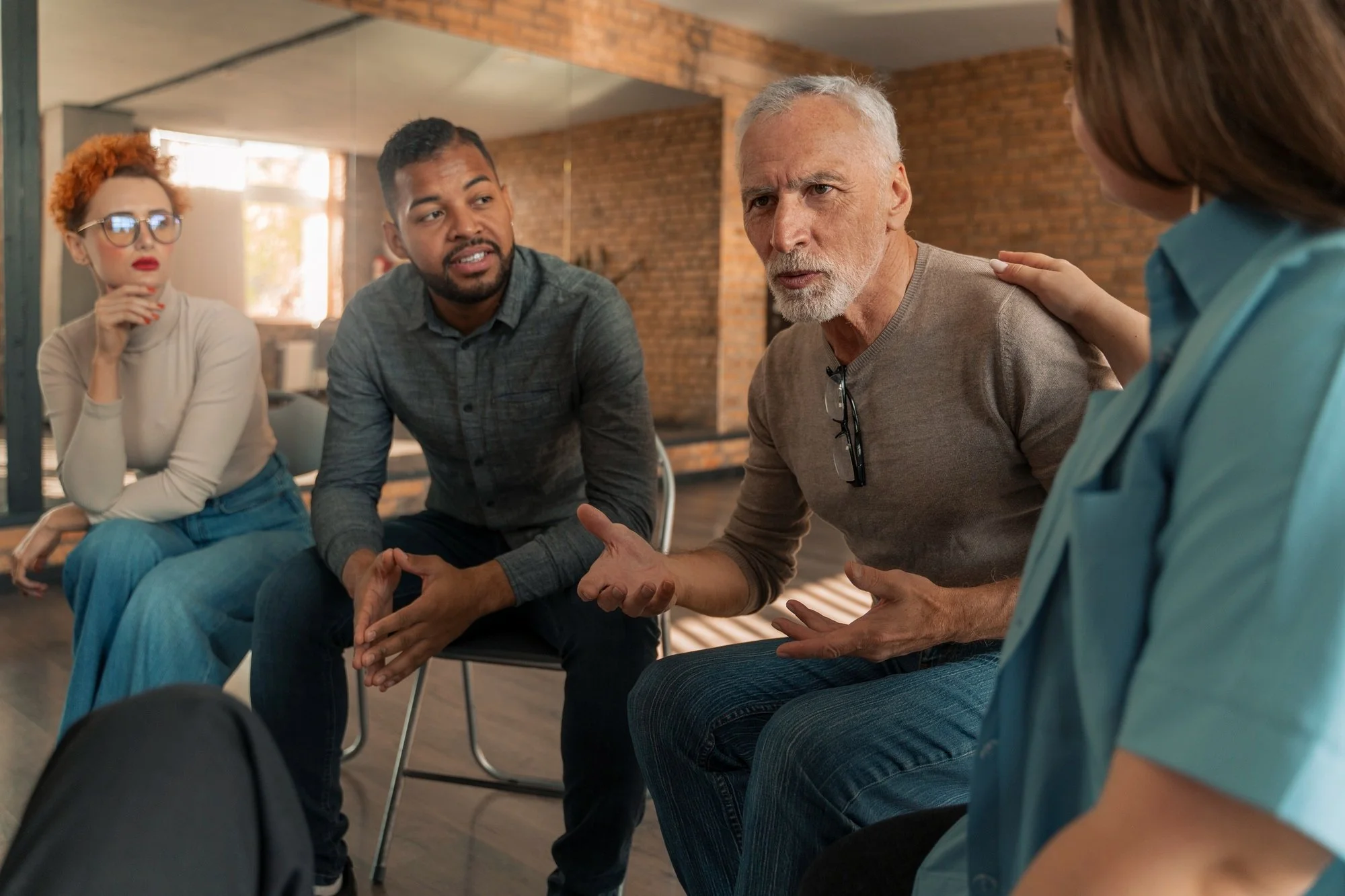 A diverse group of five people engaged in a serious conversation, seated in a room with brick walls and large windows, with some individuals actively gesturing and listening intently.