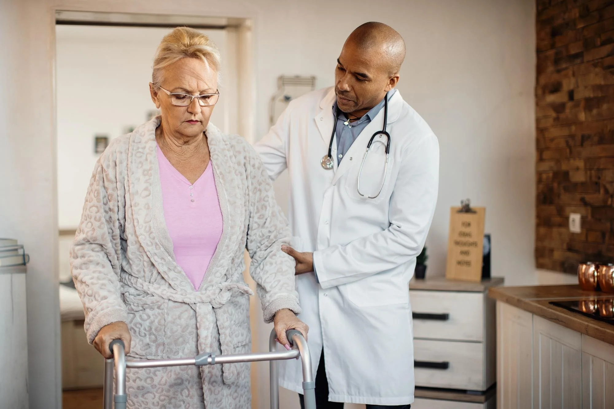 A male doctor in a white coat with a stethoscope around his neck assists an elderly woman in a pink top and patterned dressing as she uses a walker in a medical office.
