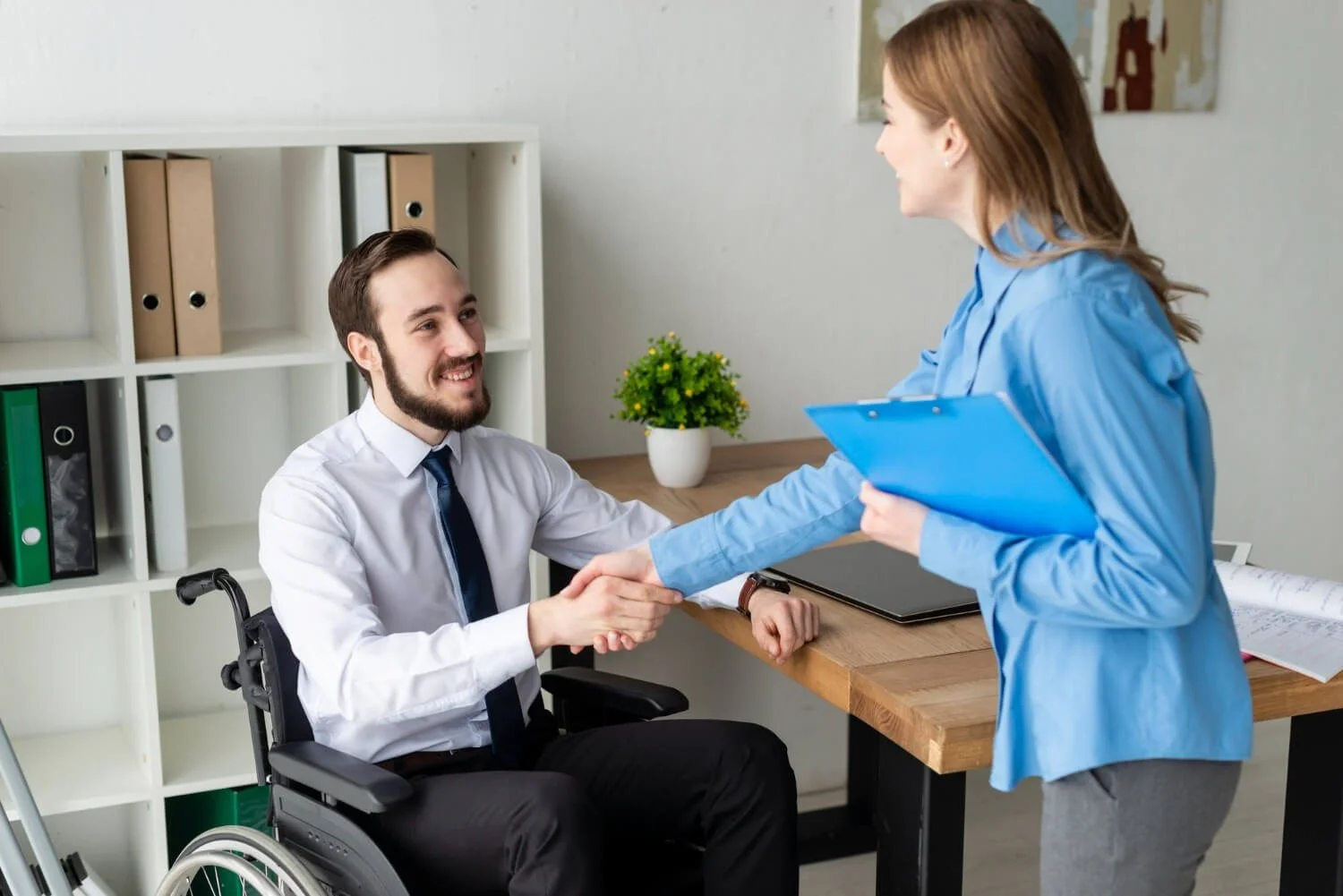 A man in a wheelchair shaking hands with a woman holding a blue clipboard in an office setting.