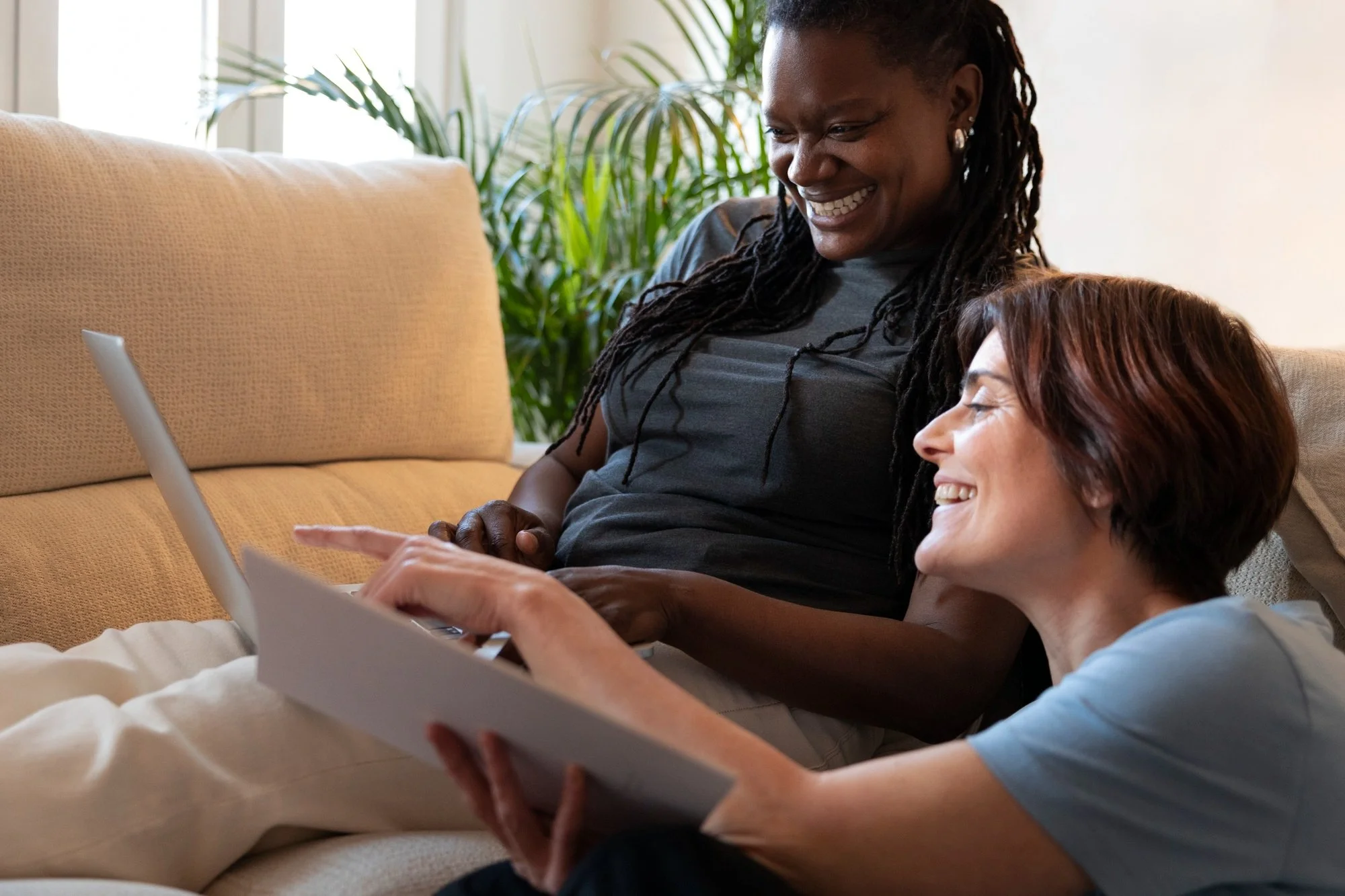 Two women, one with dark curly hair and the other with short reddish hair, sitting on a couch looking at a laptop and smiling.