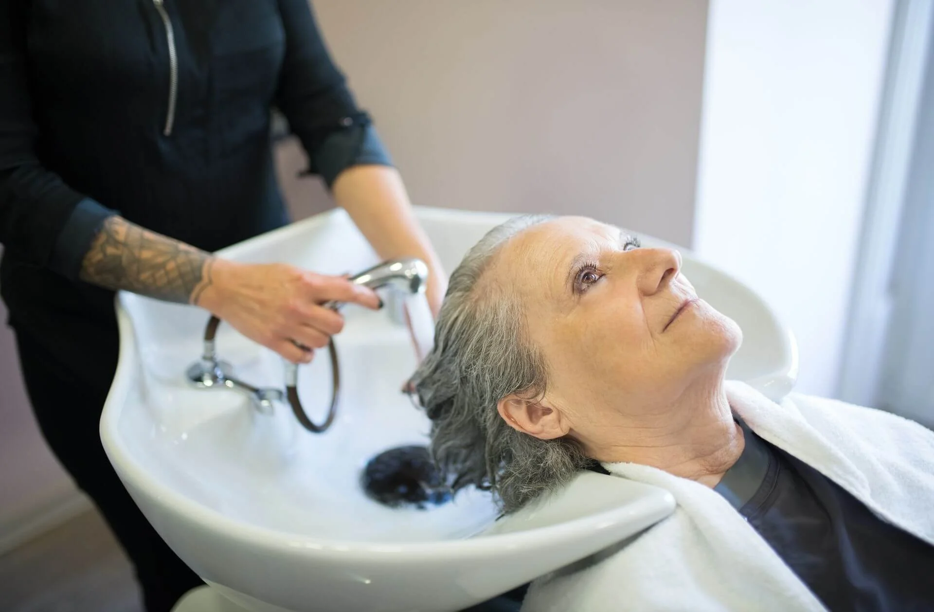 An elderly woman with gray hair receiving a hair rinse at a salon sink, while a stylist in black attire rinses her hair.