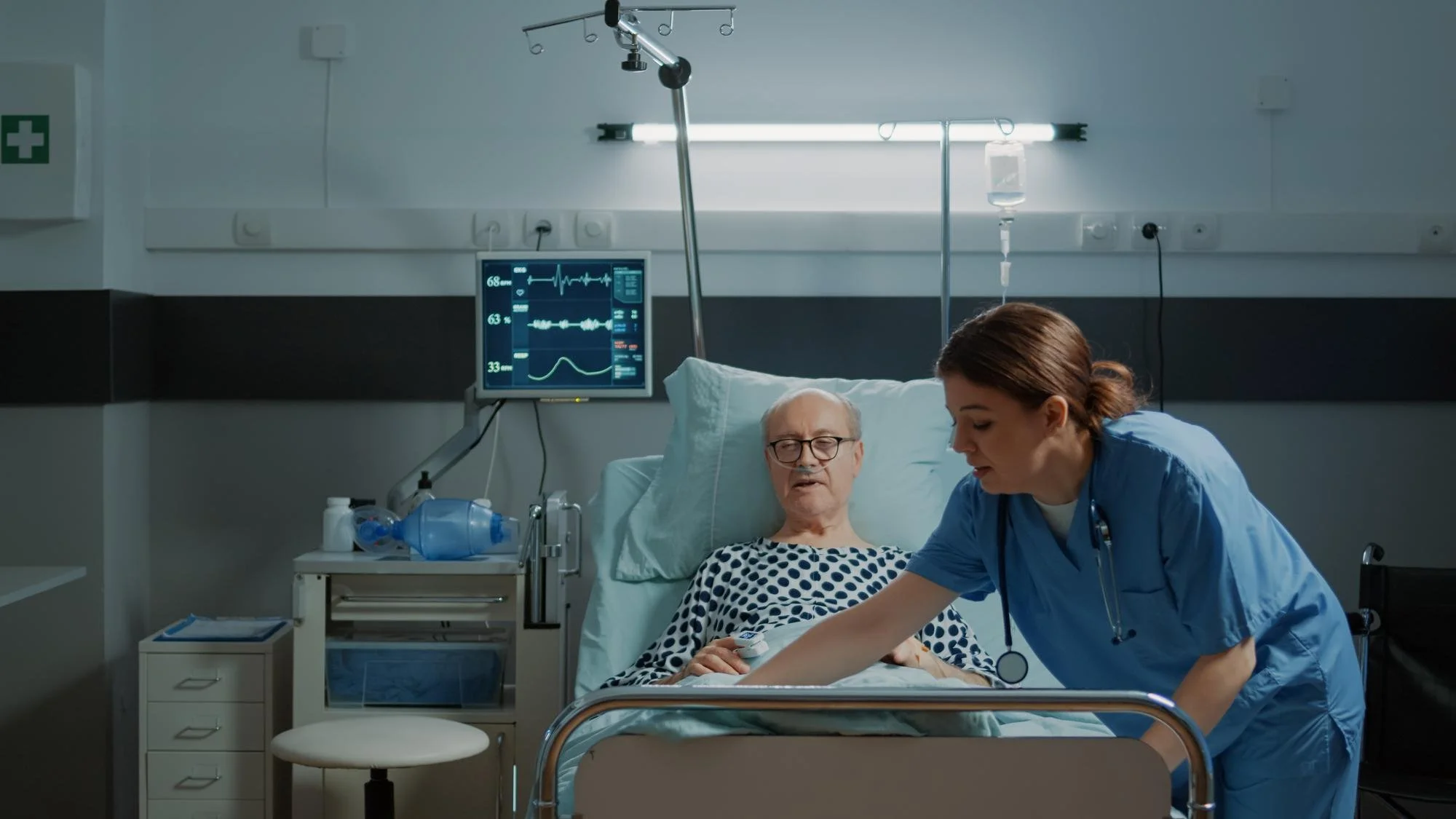 A male patient lying in a hospital bed with a nurse attending to him. The patient is elderly, wearing glasses, and a polka dot hospital gown. The nurse is in blue scrubs and is checking the patient, who appears to be conscious. Medical equipment such as a monitor displaying vital signs and an IV drip are visible in the room.