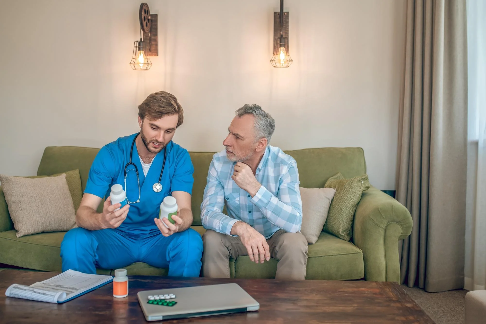 A young male healthcare worker in blue scrubs with a stethoscope around his neck shows two medicine bottles to an older man in plaid shirt seated on a green sofa in a living room. There are medication bottles, a notebook, and a laptop on the table in front of them.