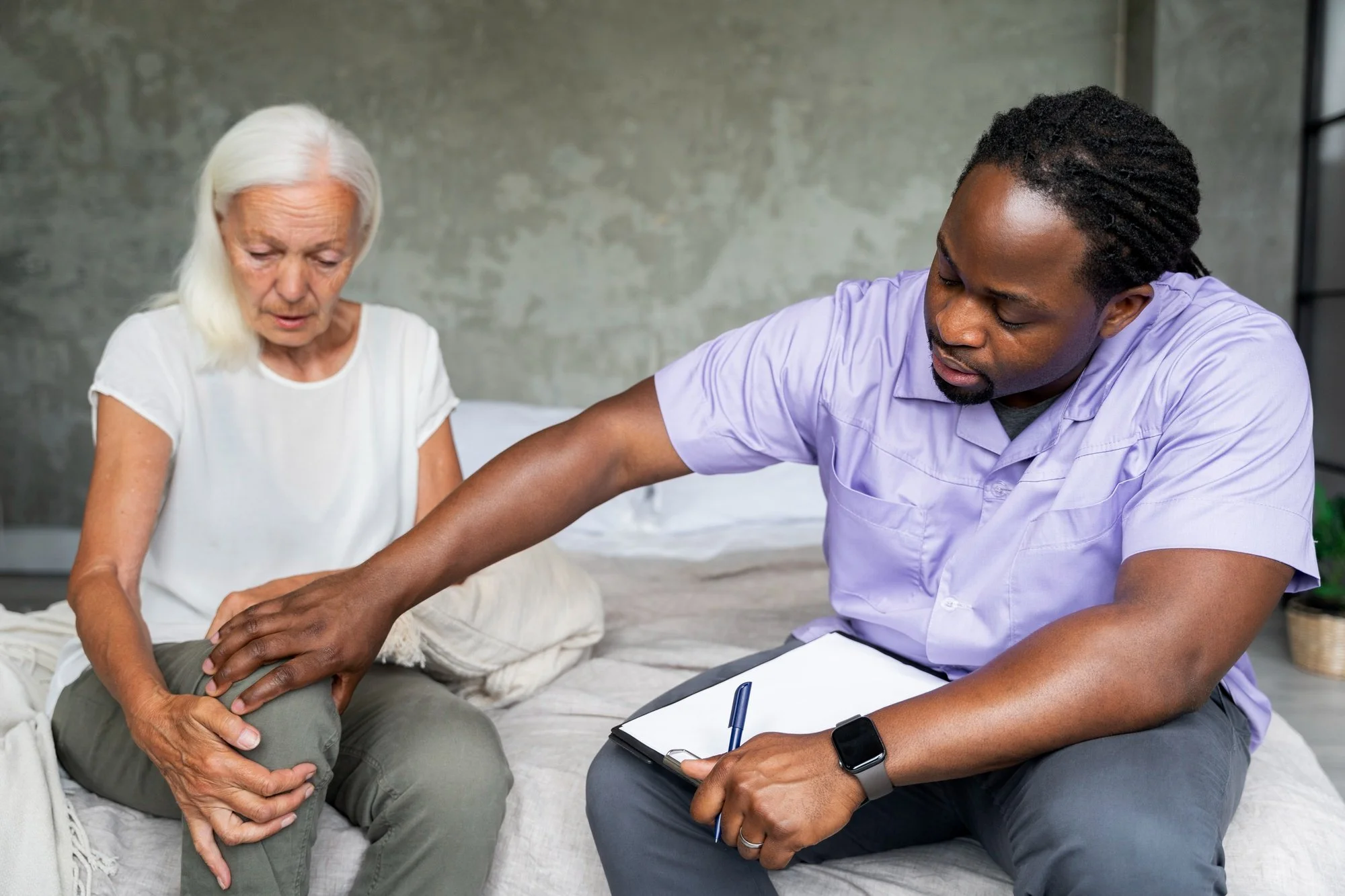 A healthcare professional examining an elderly woman's knee during a consultation in a clinical setting.