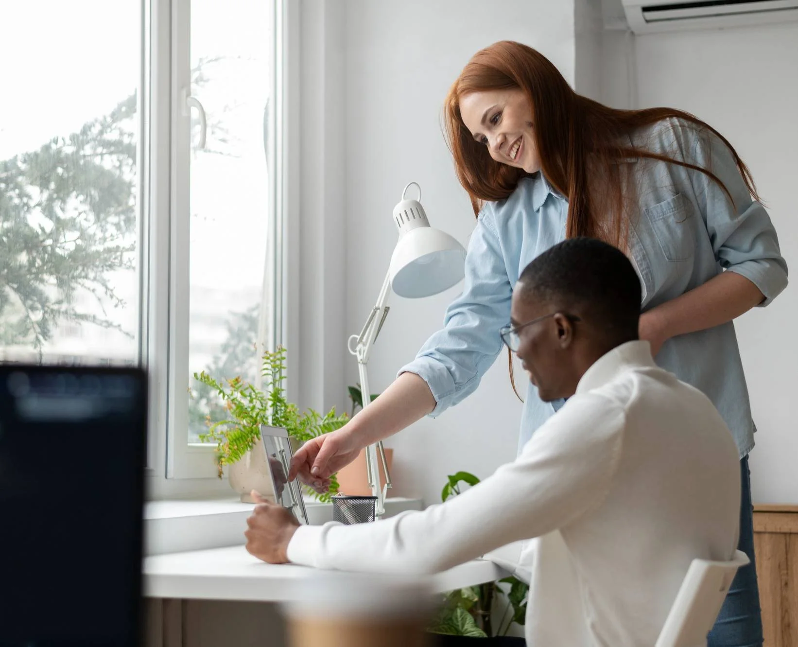 A woman with long red hair smiling and pointing at a tablet screen while a man with dark hair and glasses sits at a desk and looks at the tablet. They are in a well-lit room with a large window, houseplants, and a desk lamp.