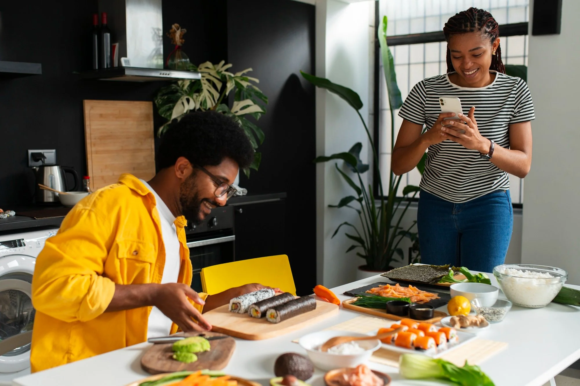 A woman with braided hair in a striped shirt smiling while looking at her phone, standing in a modern kitchen. Sitting at the counter, a man with glasses and a yellow jacket is preparing sushi on a cutting board, surrounded by various ingredients including sushi rolls, vegetables, and herbs.