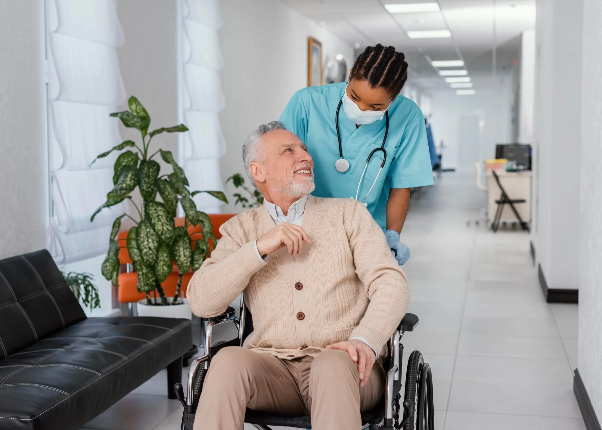 An elderly man in a beige cardigan is sitting in a wheelchair in a hospital corridor. A nurse wearing a blue medical uniform and a face mask is attending to him and looking at his face. There are white walls, a plant, and some chairs in the background.