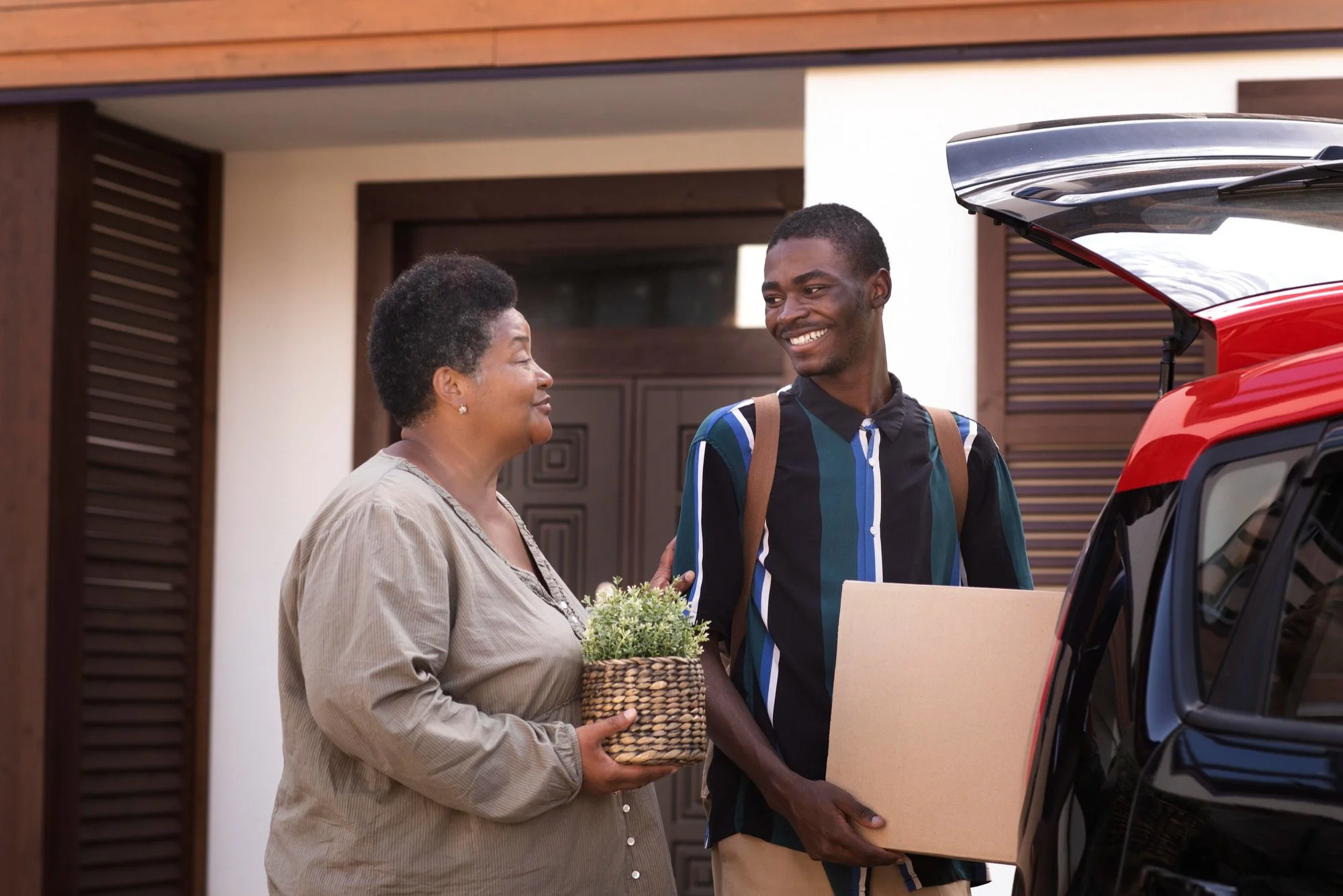 An elderly woman and a young man smiling at each other as they unload a car with a cardboard box and a flower basket outside a house.