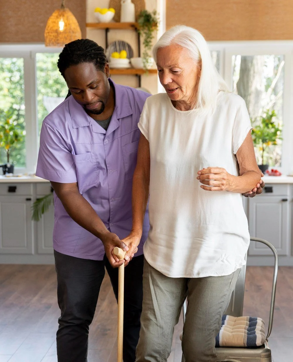 A caregiver assists an elderly woman walking with a cane in a bright kitchen.