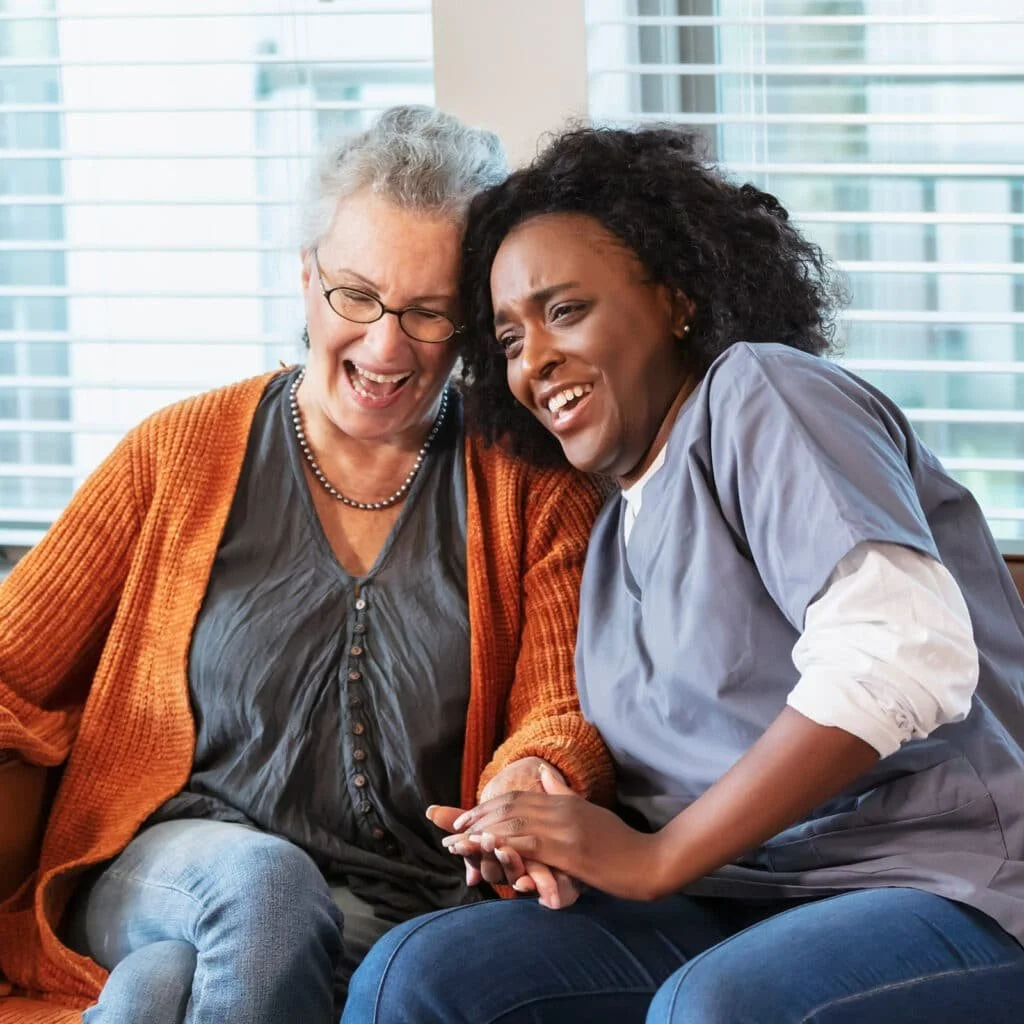 An elderly woman and a younger woman are sitting close, smiling, and holding hands in a well-lit room with large windows and blinds.