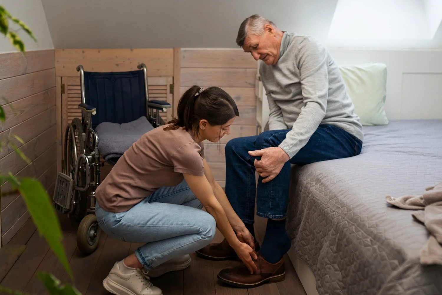 A young woman helping an elderly man put on his shoes in a bedroom, with a wheelchair nearby.