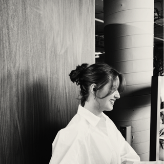 A woman with dark hair tied in a bun, wearing a white blouse, smiling and looking downward, standing indoors near a textured wall and modern architectural columns.