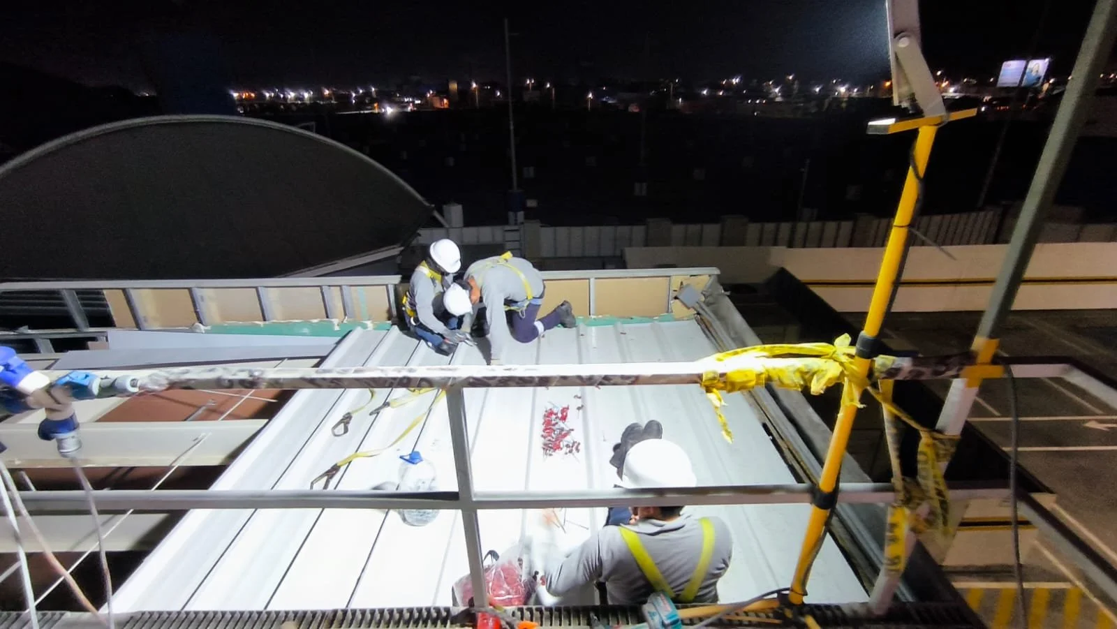 Trabajadores en la azotea de un edificio realizando mantenimiento durante la noche, usando equipo de seguridad y casco.