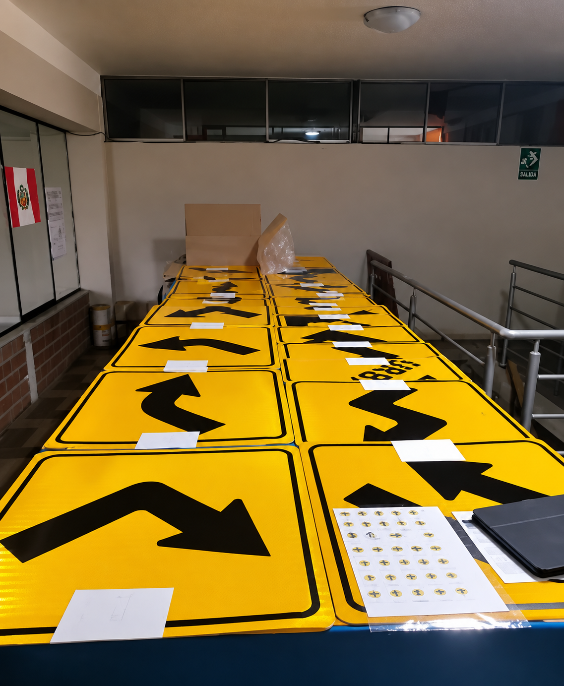 Mesas con señalizaciones amarillas y flechas negras para tránsito o dirección, posibles instrucciones viales, en un espacio interior con ventanas y una señal de salida en la pared.