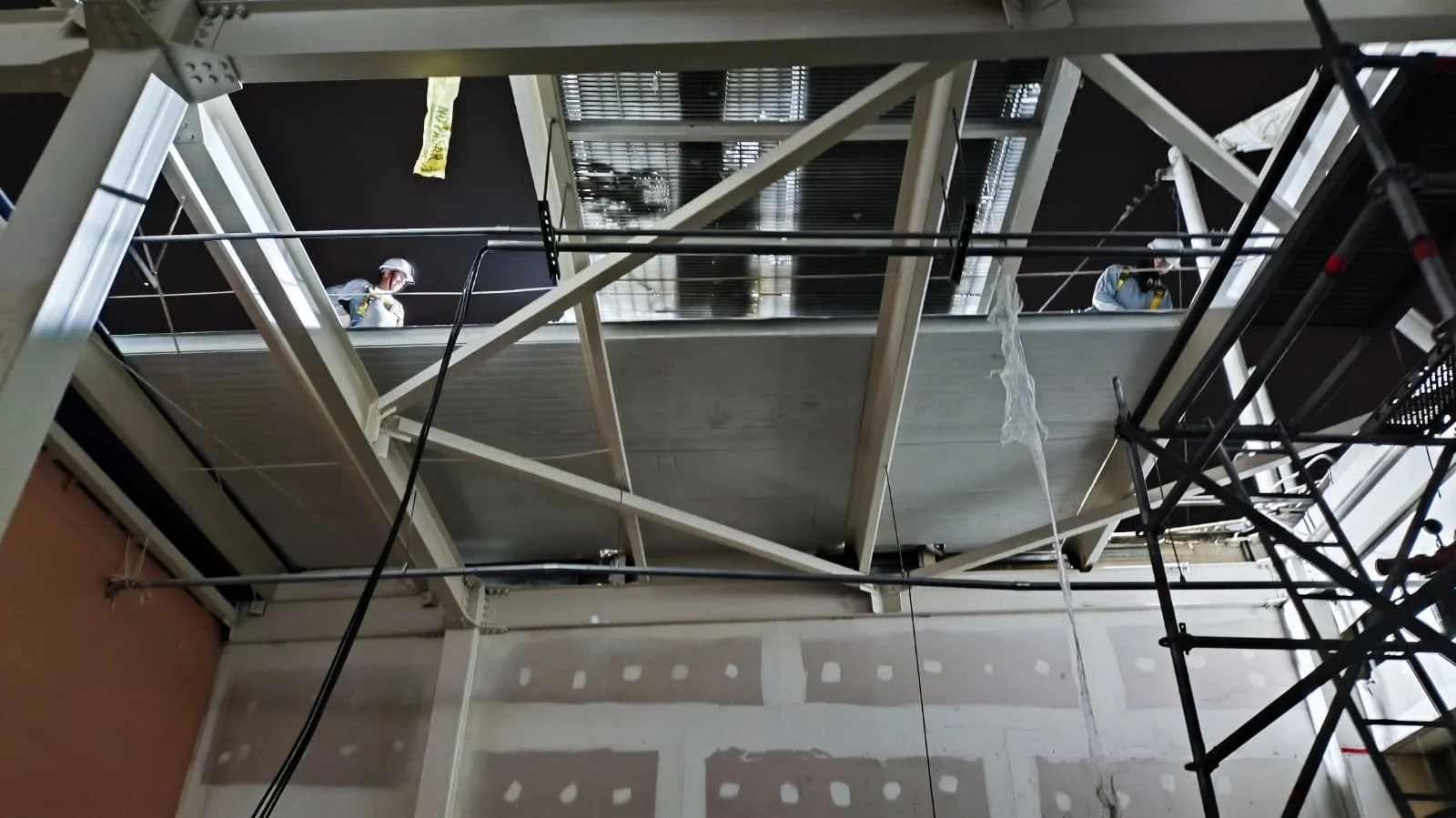 Trabajadores en un andamio en construcción en interiores, con paredes en yeso y escaleras metálicas.
