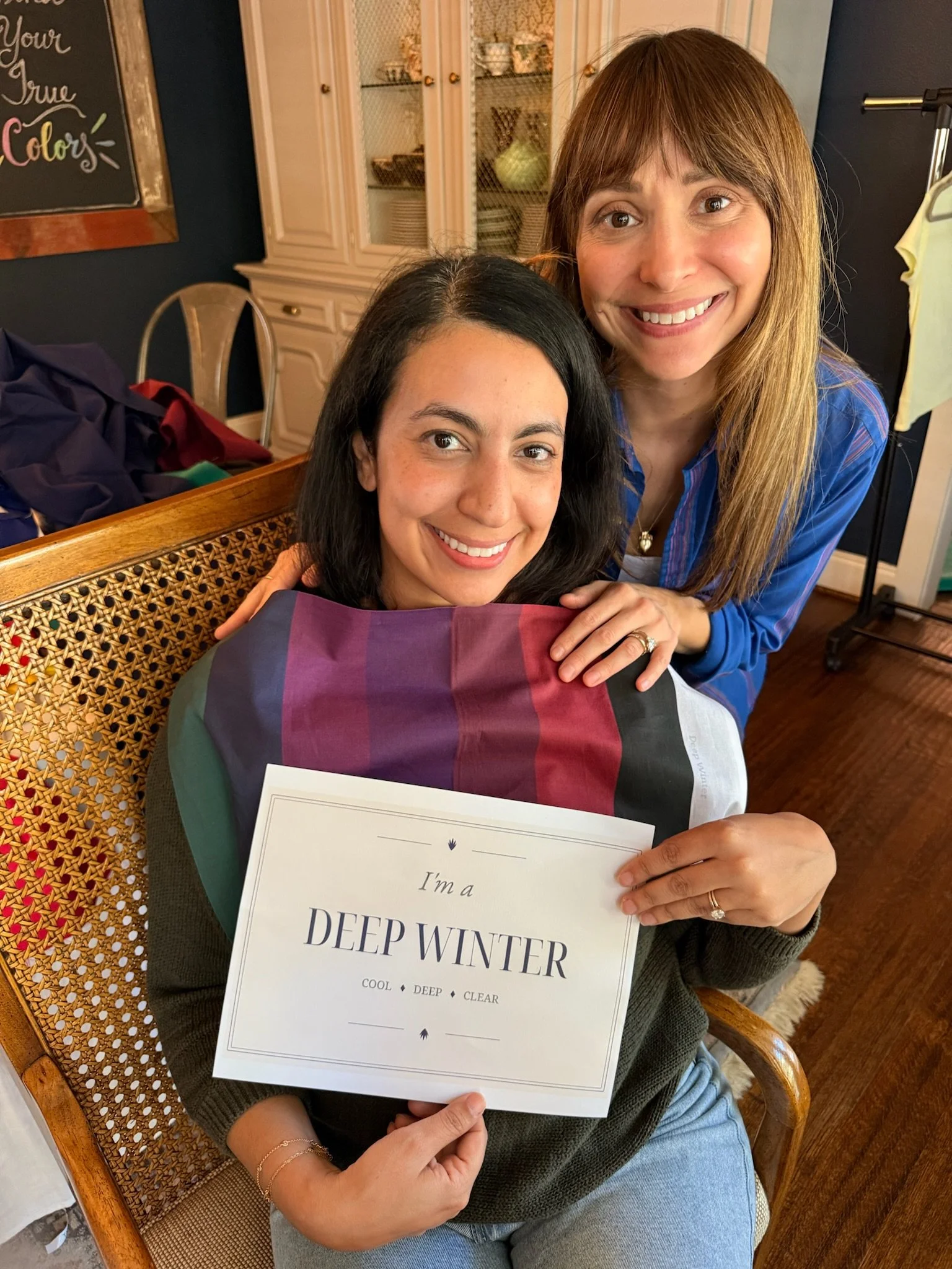 Two women smiling indoors, one sitting, holding a sign that reads 'I'm a deep winter. Cool, deep, clear.' The other woman is standing behind her. There is a wooden table with clothes and a chalkboard in the background.