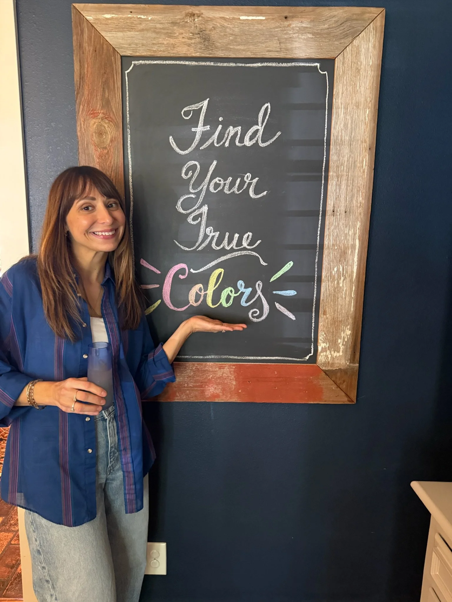 A woman holding a small chalkboard with the words 'Find Your True Colors' written on it. She is standing next to a larger chalkboard with the same message. She has shoulder-length brown hair and is smiling.