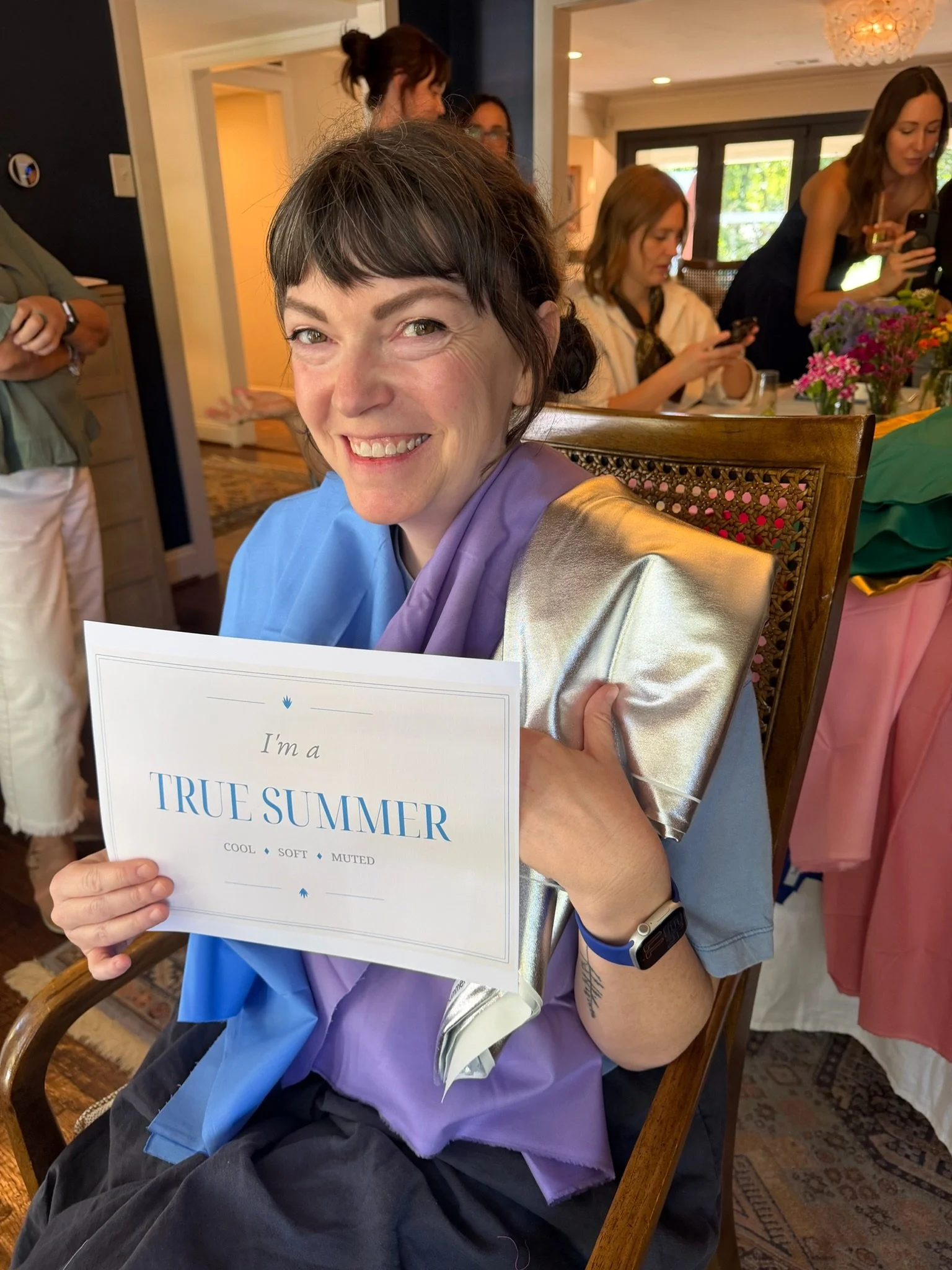 A smiling woman holding a card that reads 'I'm a TRIPLE SUMMER' in a room with other people who are engaged with their phones and conversations, with floral decorations on the table behind her.