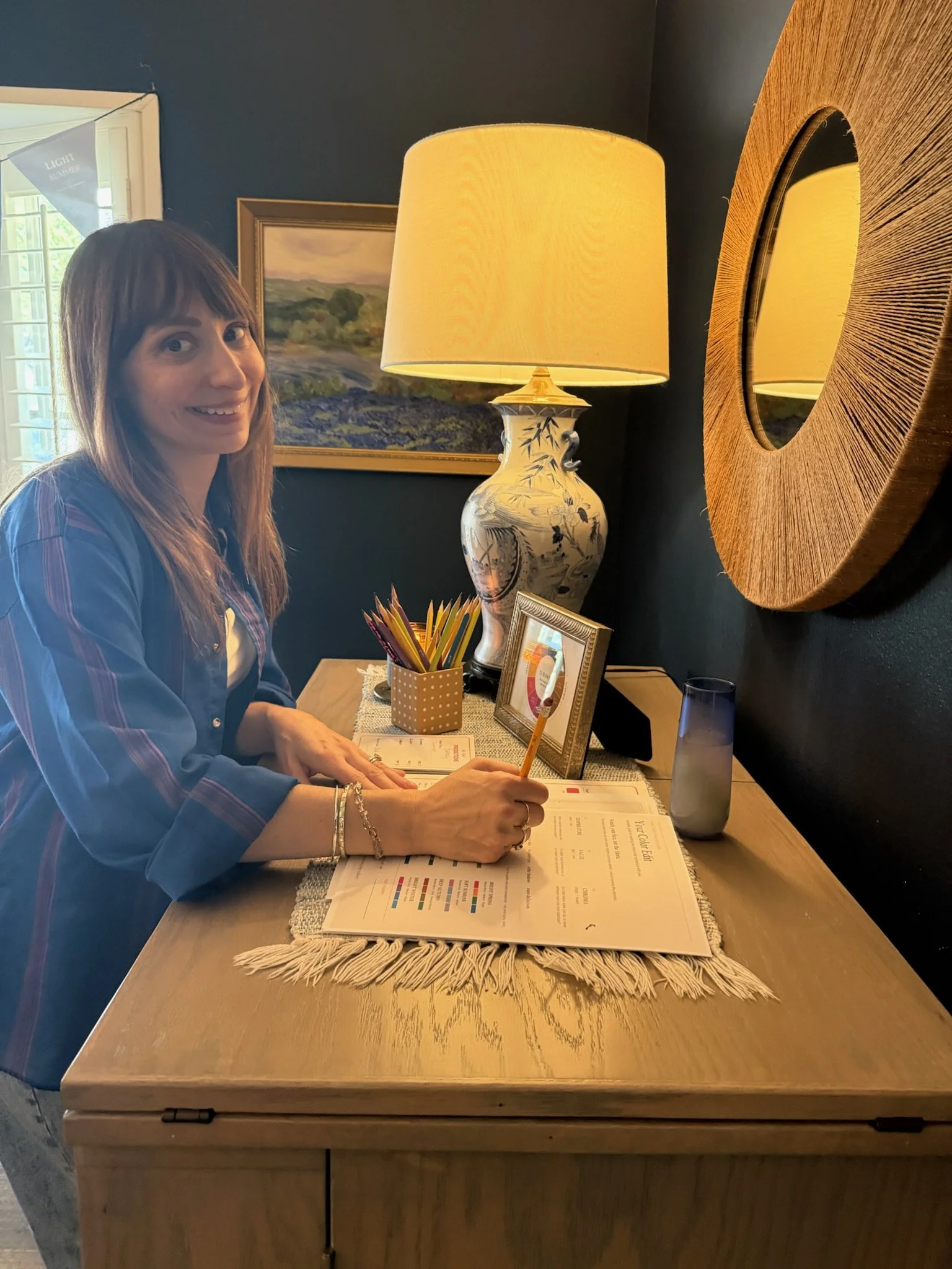 A woman with long hair smiling and writing in a menu at a dining table. The table has a beige table runner, a small container with colored pencils, a framed picture, a tall grey glass, and a large decorative lamp with a yellow shade. The background includes a dark wall, a framed landscape painting, a round mirror, and a window with blinds.