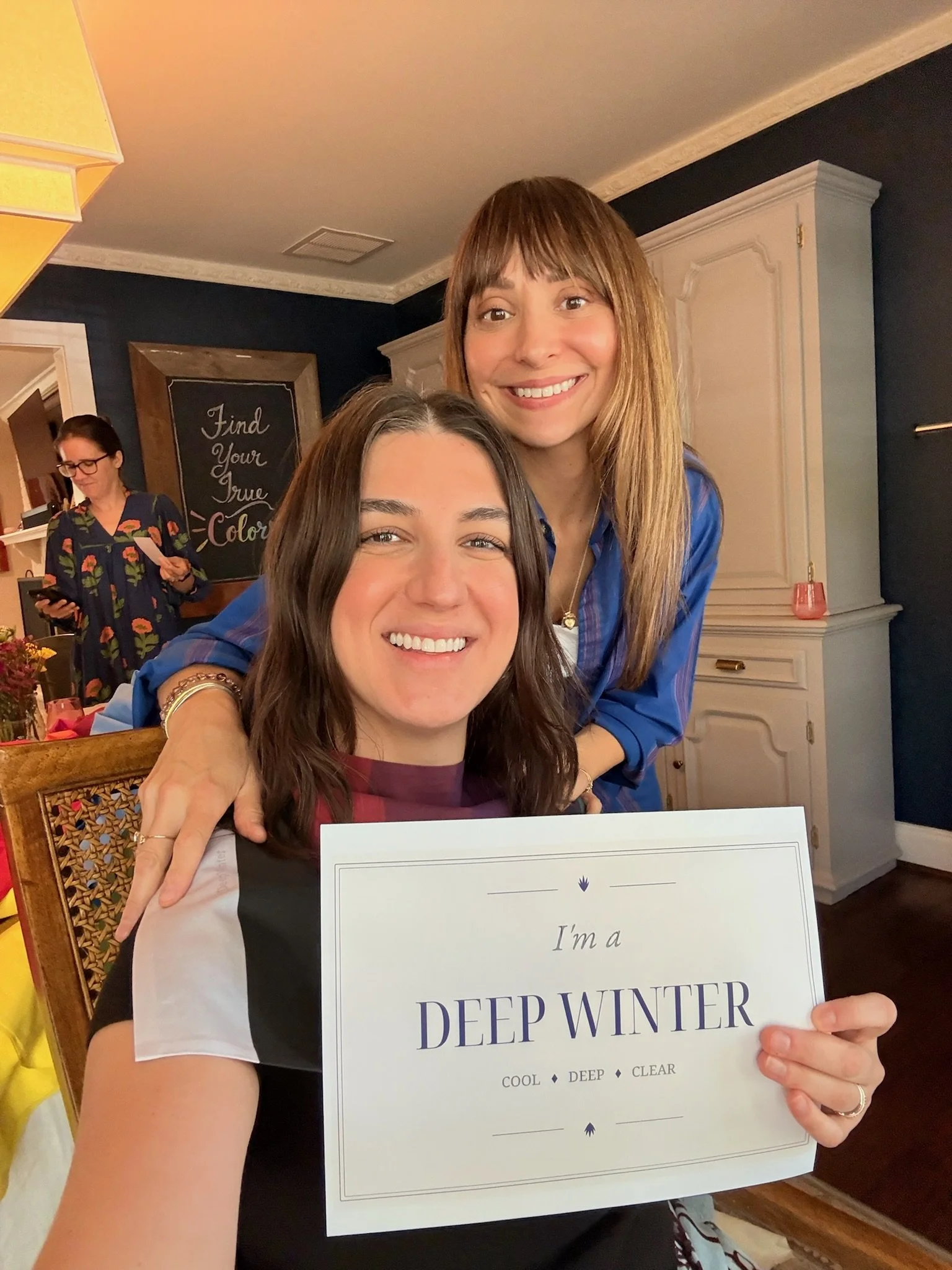 Two women smiling, one holding a certificate that says 'I'm a DEEP WINTER,' in a cozy room with dark blue walls, white trim, and a chalkboard in the background with the words 'Find Your True Color.'
