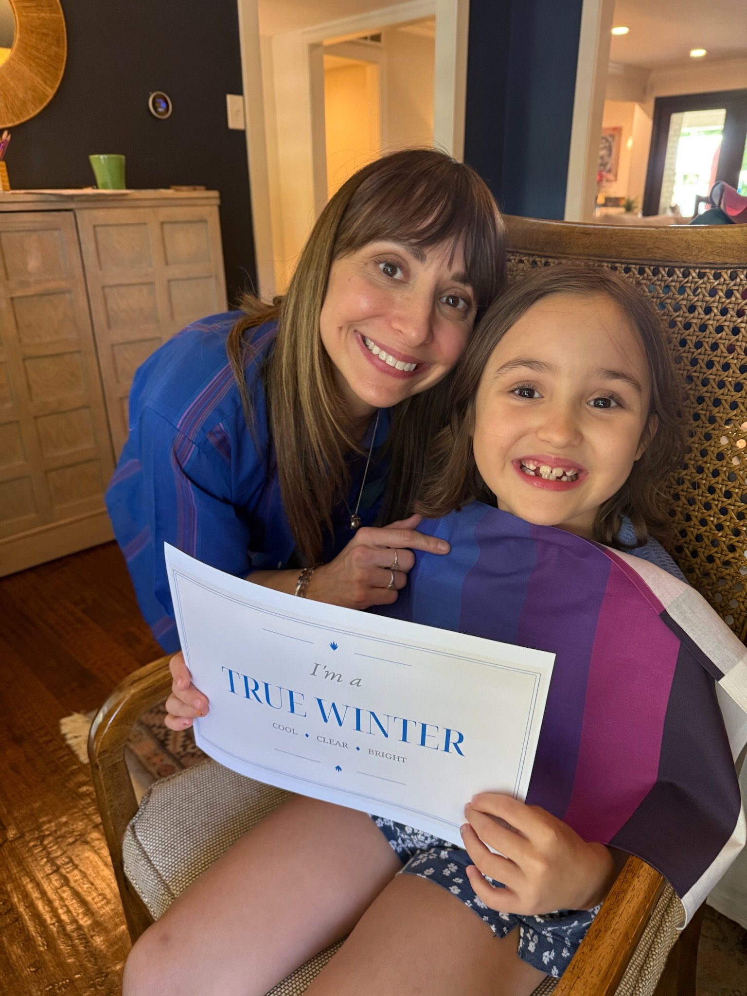 A woman and a young girl sitting on a chair, smiling at the camera. The girl has missing teeth, and the woman is holding a sign that reads 'I'm a TRUE WINTER'.
