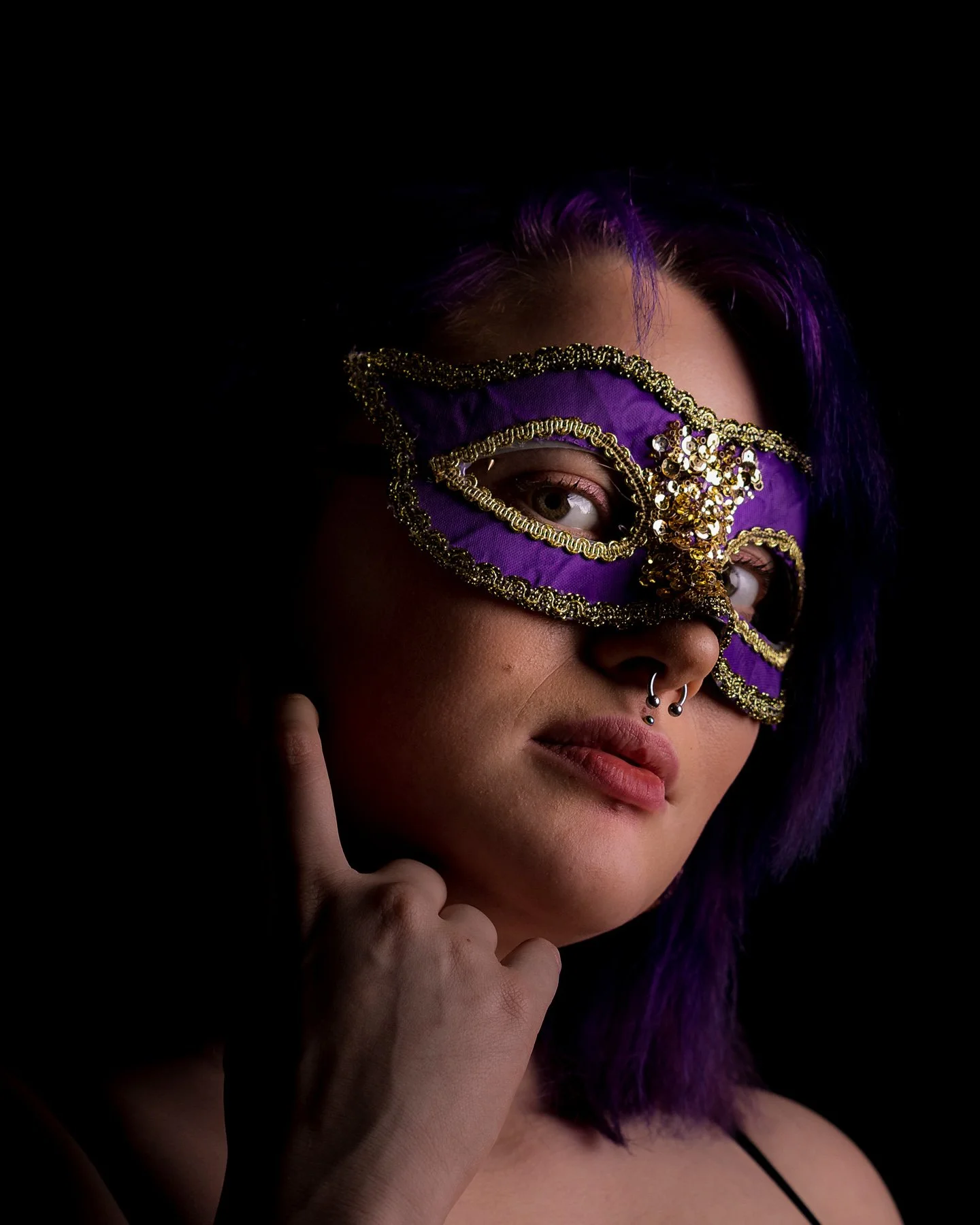 En ung kvinna med lila hår bär en ornately dekorerad purple mask som täcker ögonen, mot en mörk bakgrund.