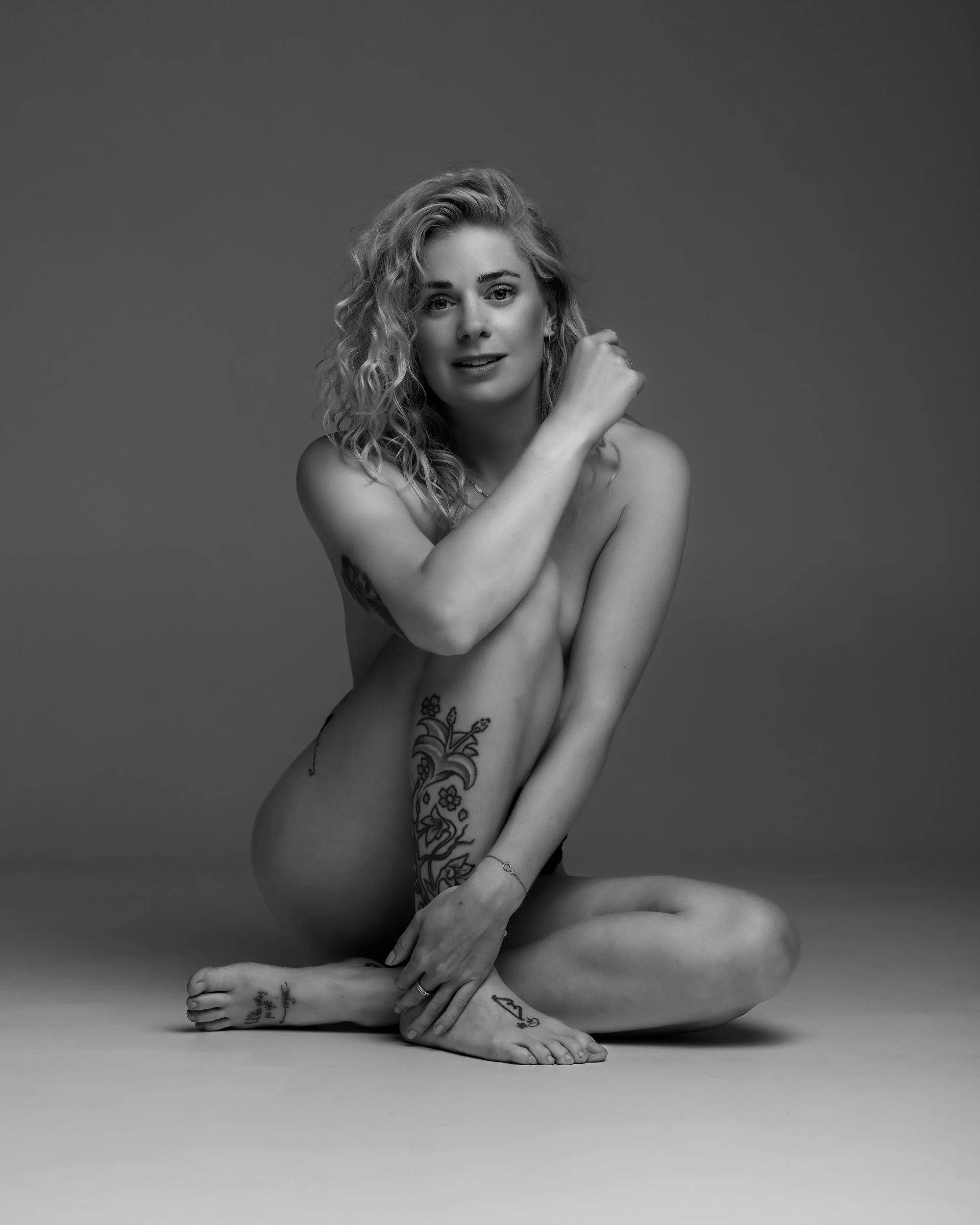 En ung kvinna sitter tvärs i en studio, naken med tatueringar på armar, ben och kropp, och har långt, lockigt hår. Hon ler lätt och tittar mot kameran.