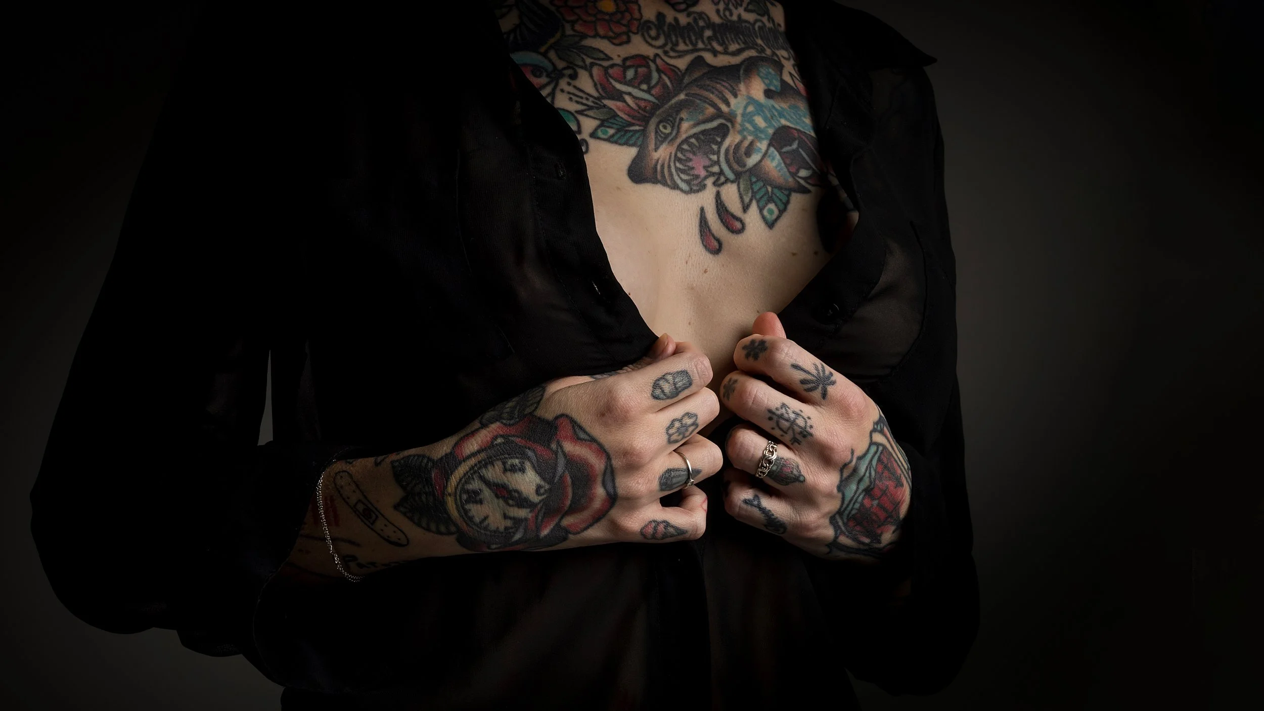 En person med många tatueringar, inklusive ett leopardhuvud, som håller på att öppna en svart skjorta eller jacka, mot en mörk bakgrund.