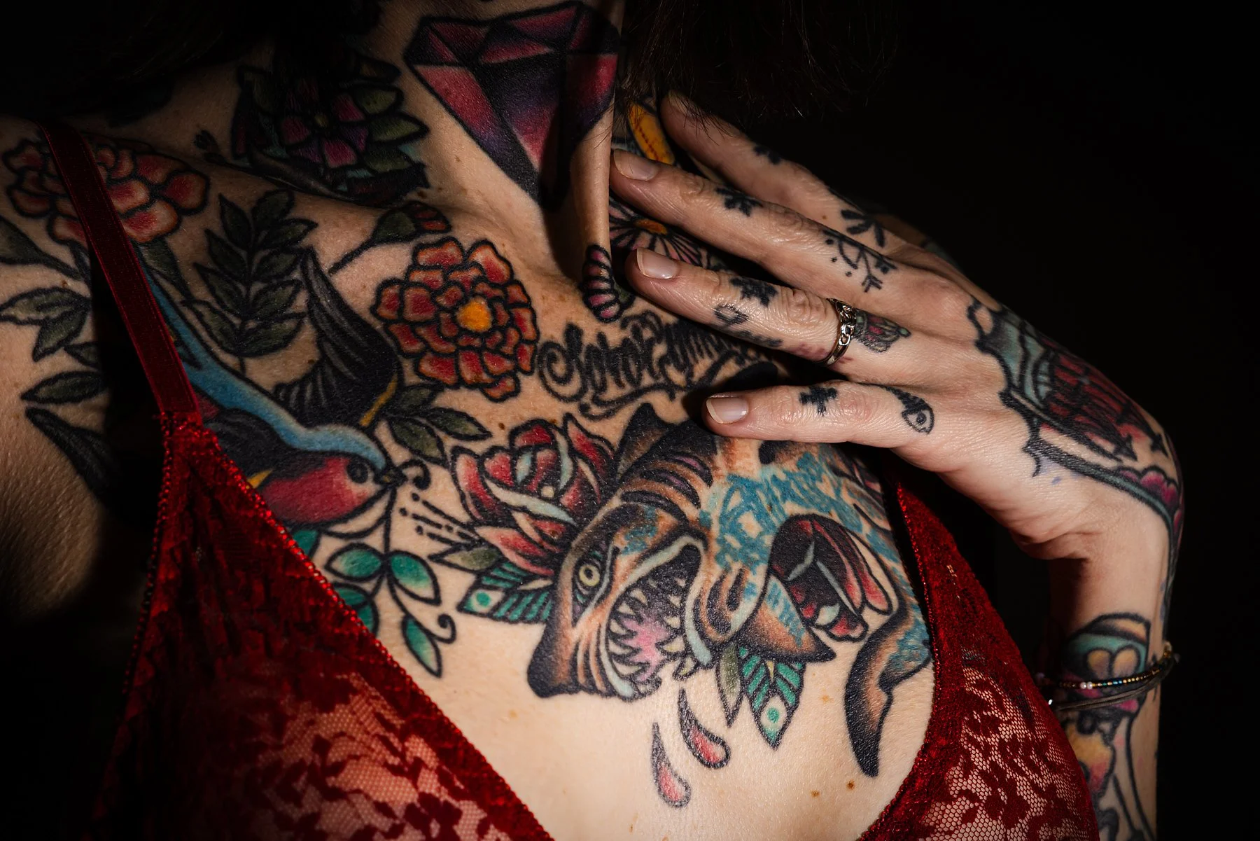 En person med många tatueringar på hals, axlar och bröst, klädd i en röda spetsklänning. Personen har fingrar med ringar och visar en hand mot halsen. Tatueringarna inkluderar blommor, djur och text.