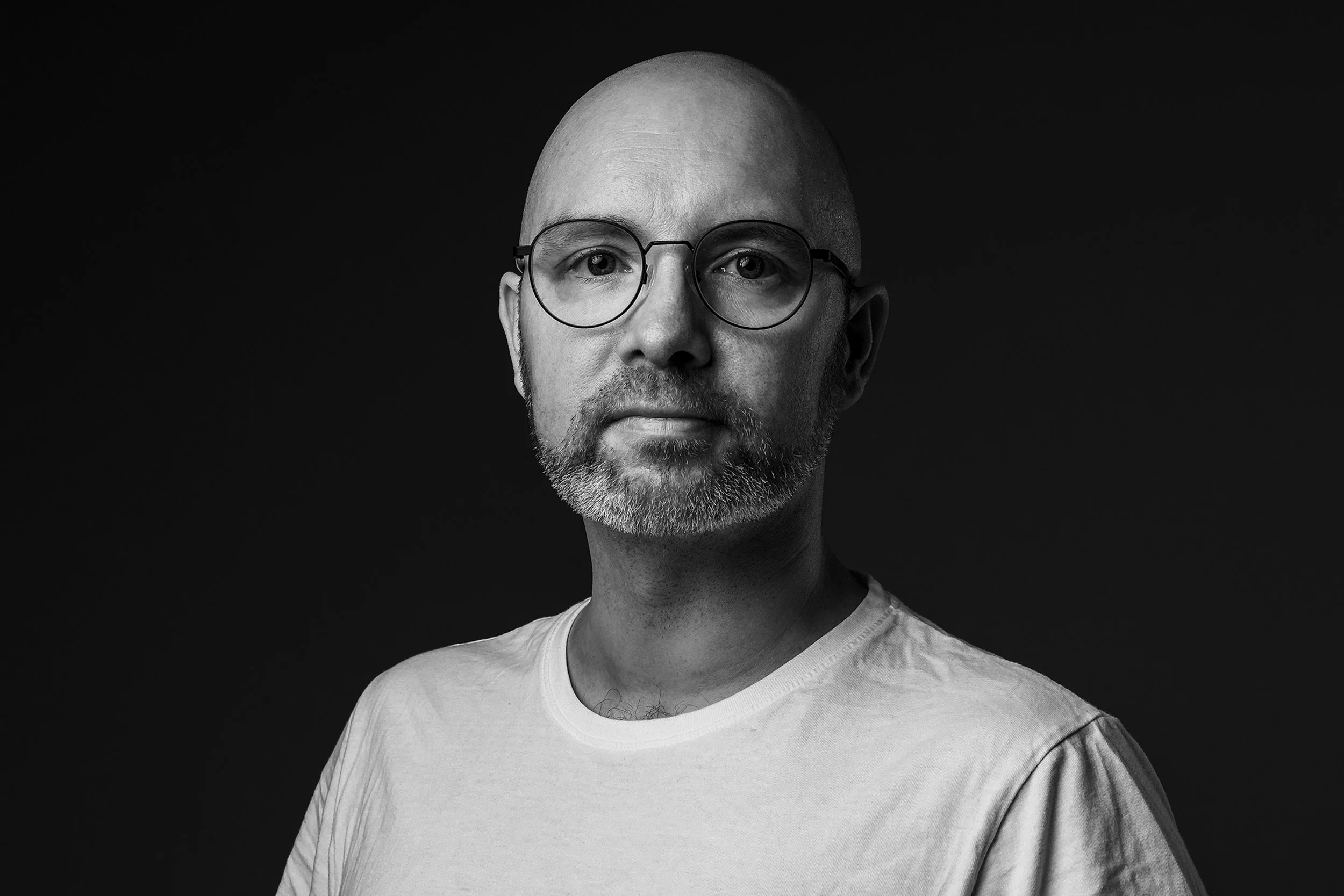 Svartvit bild av en man med glasögon, skägg och mustasch, som bär en vit T-shirt, mot en mörk bakgrund.