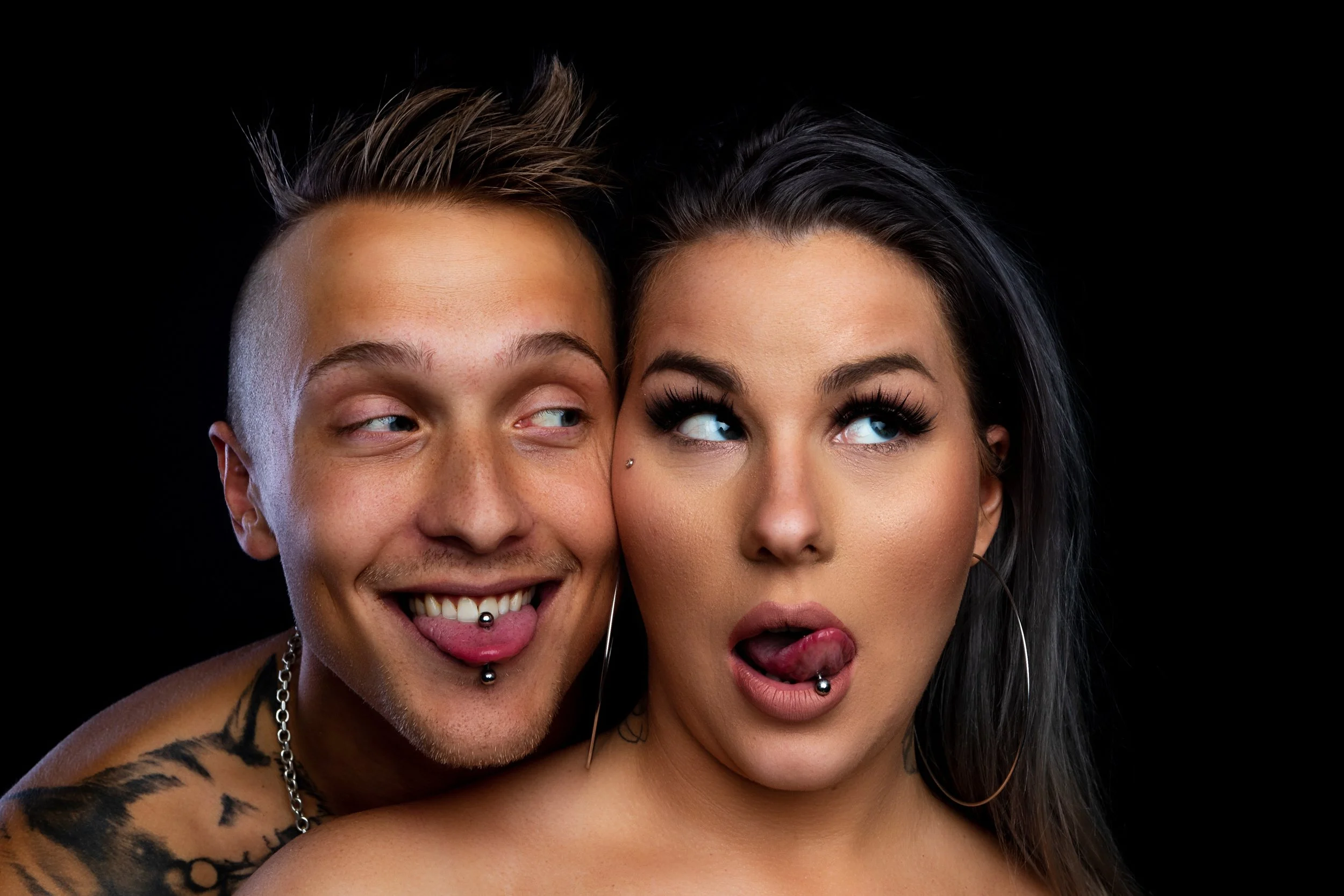 Två personer nära varandra, en med kort hår och piercing på tungan, den andra med långt mörkt hår och piercing på läppen, gör ansiktsuttryck. Bakgrund svart.
