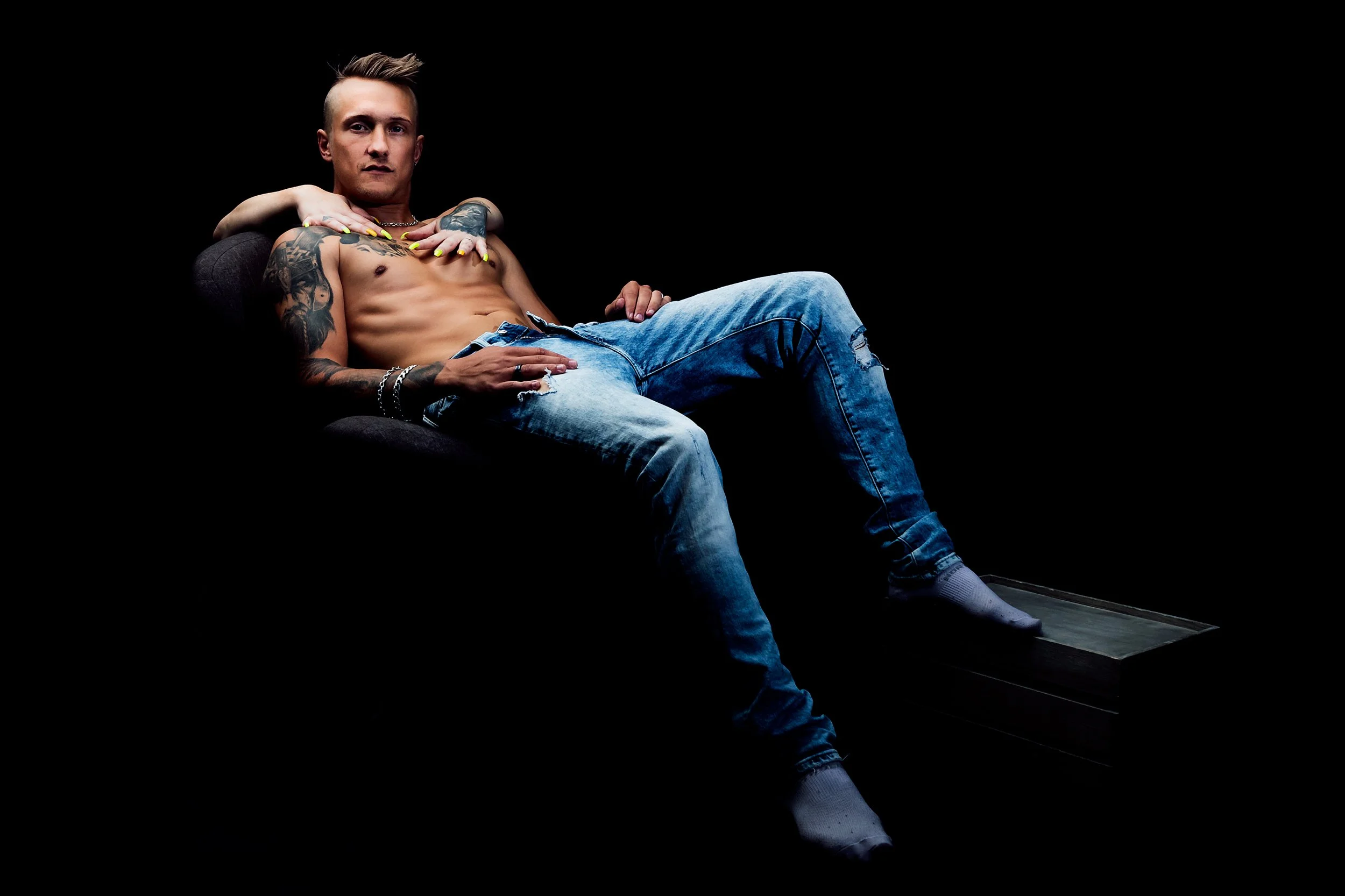 En ung man med tatueringar, blå jeans och vita strumpor sitter på en svart stol mot en svart bakgrund.