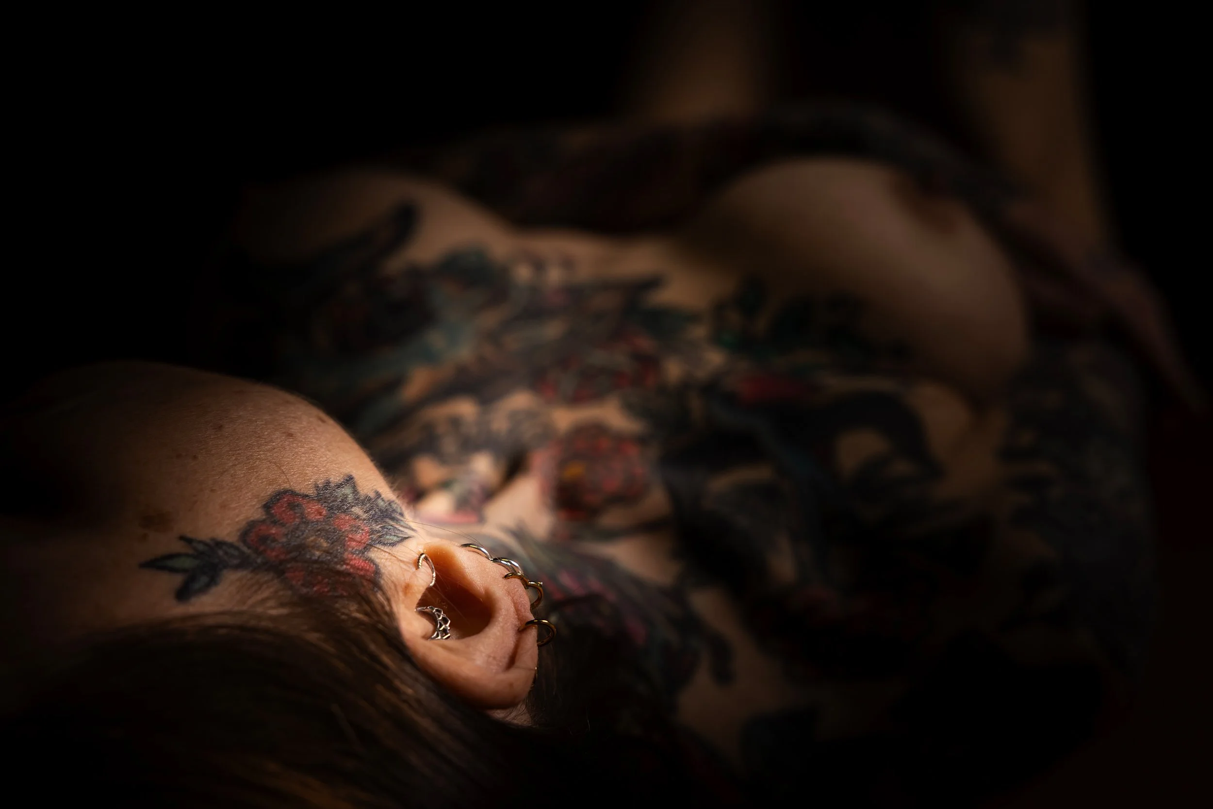 En person med tatueringar och piercingar ligger på rygg, omgiven av mörkt omgänget. Skuggor faller över personen och bakgrunden är svart.