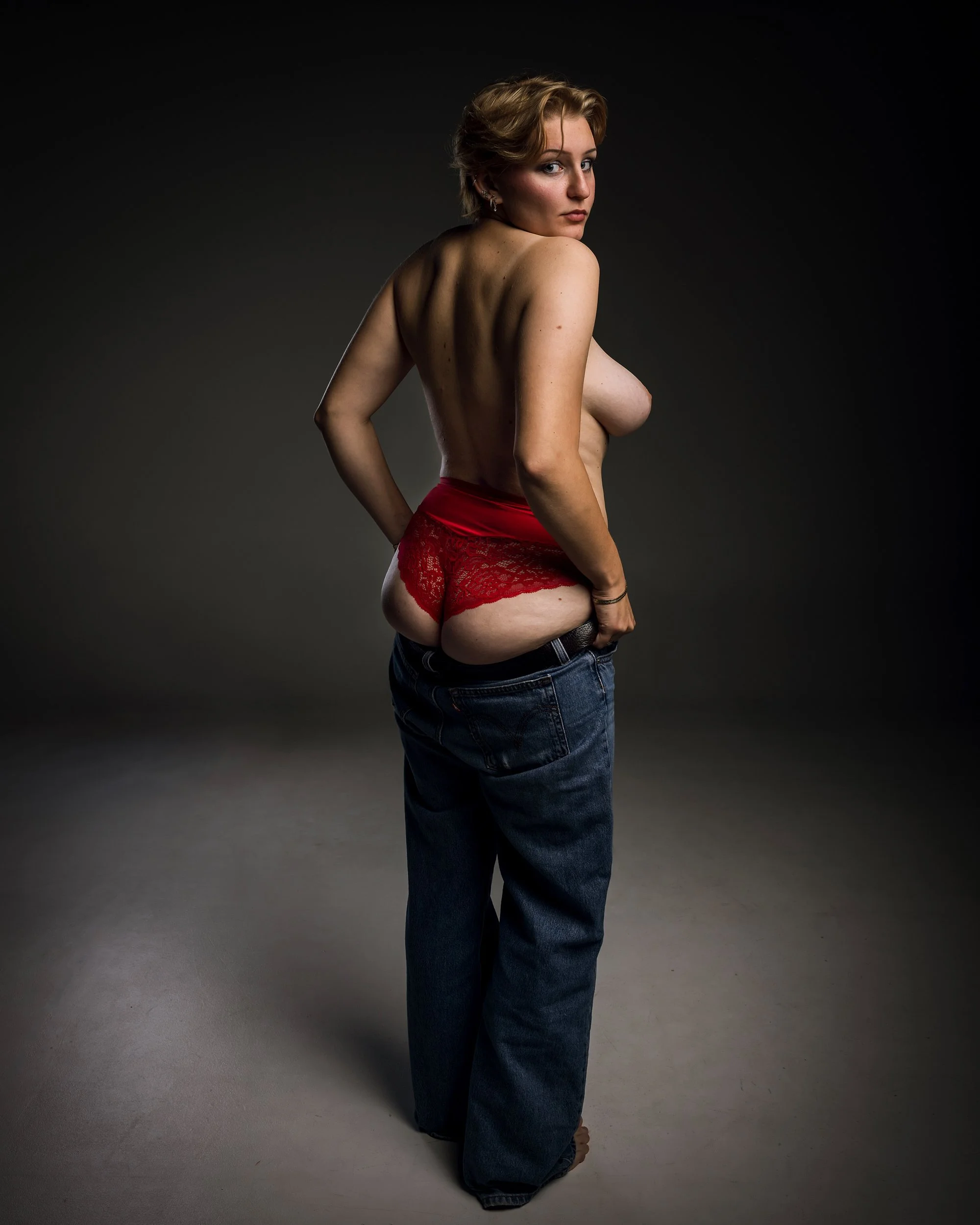 En person med blont hår bär endast röda underkläder och mörka jeans medan hon står mot en mörk bakgrund.