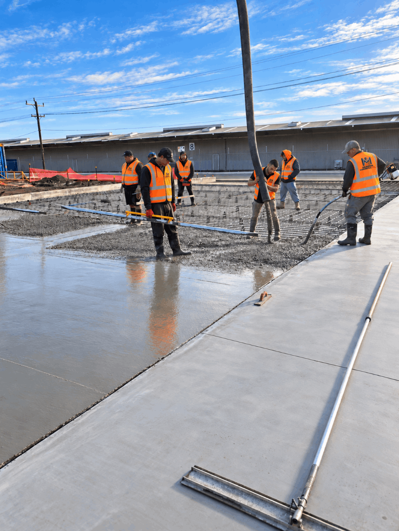Construction workers wearing orange safety vests pouring and leveling concrete on a building site under a blue sky.