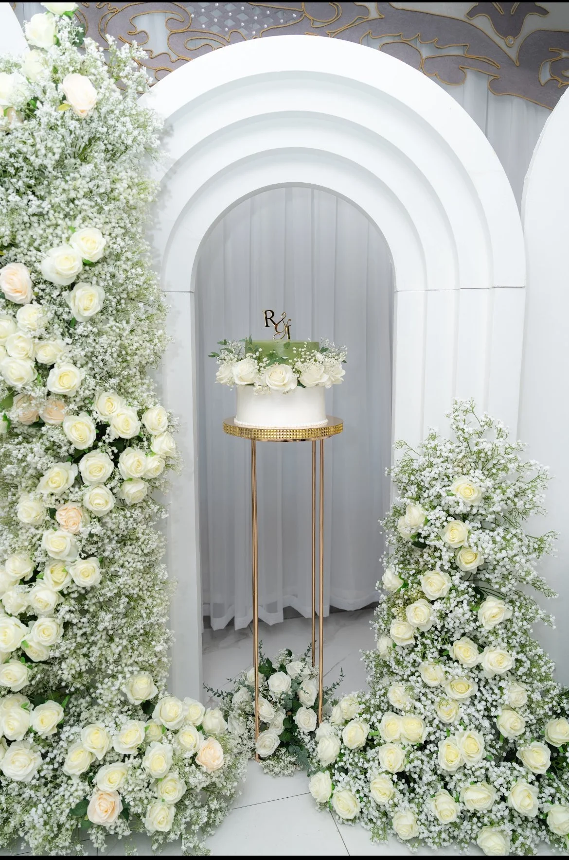 A wedding or special event setup featuring a white cake decorated with flowers, placed on a tall gold stand, surrounded by arrangements of white roses and baby's breath flowers on both sides, with a white arched backdrop.