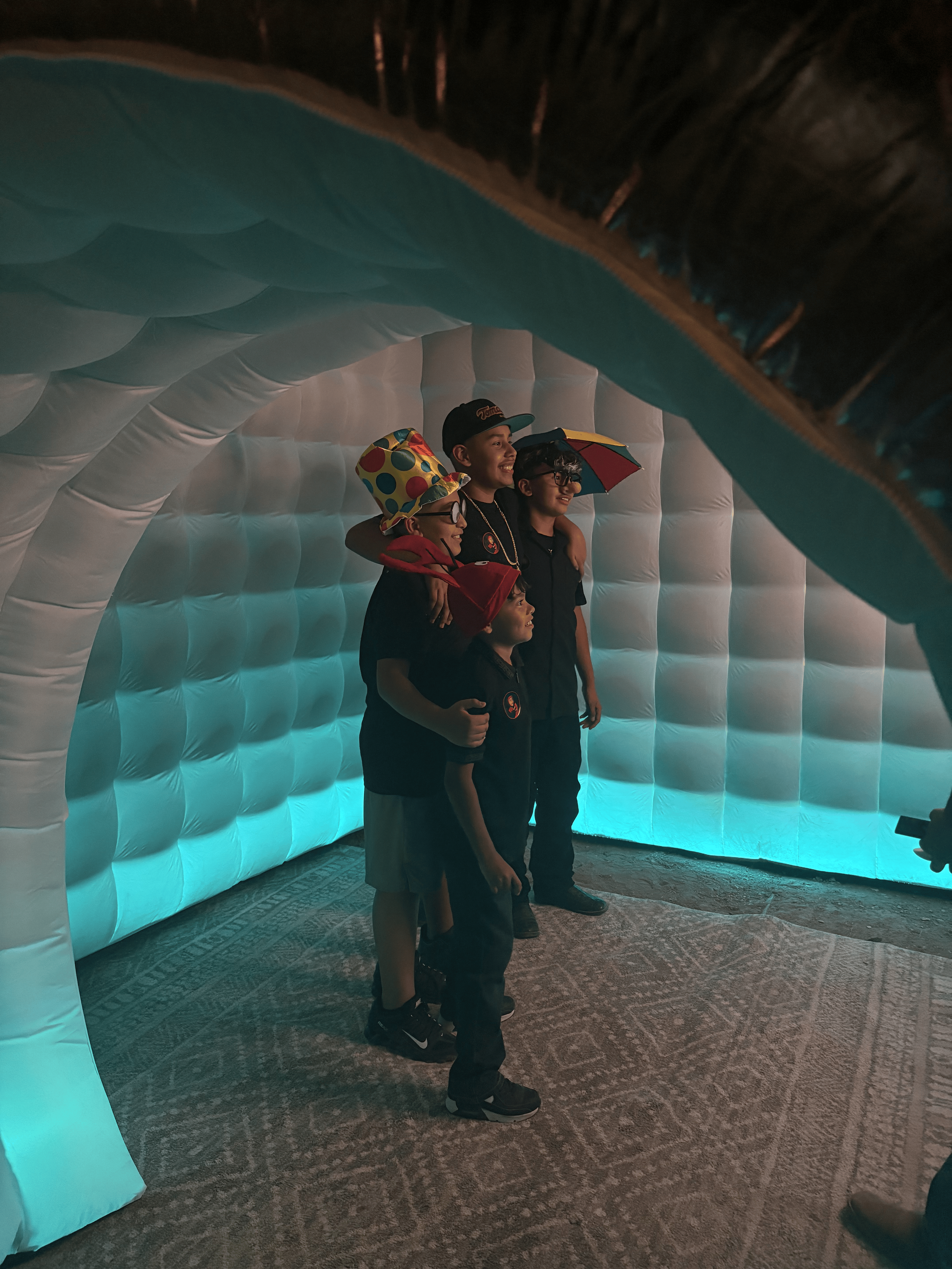 Four boys posing for a photo inside an inflatable photo booth with illuminated walls. Two of the boys are wearing glasses, and one is holding a colorful umbrella. They are smiling and standing close together.