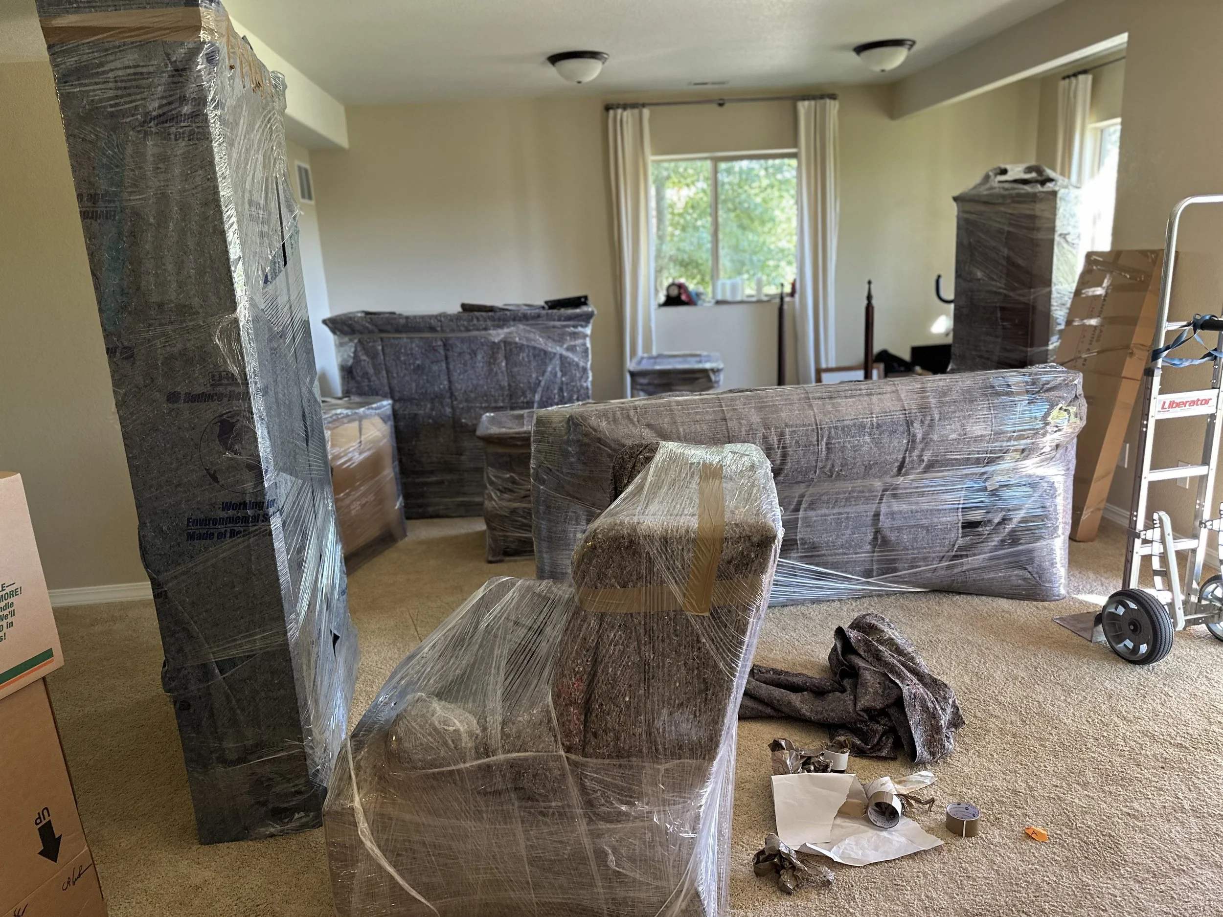 Living room with furniture wrapped in protective plastic for moving, including a sofa and chair. Cardboard boxes and moving supplies scattered around, with a hand truck visible.