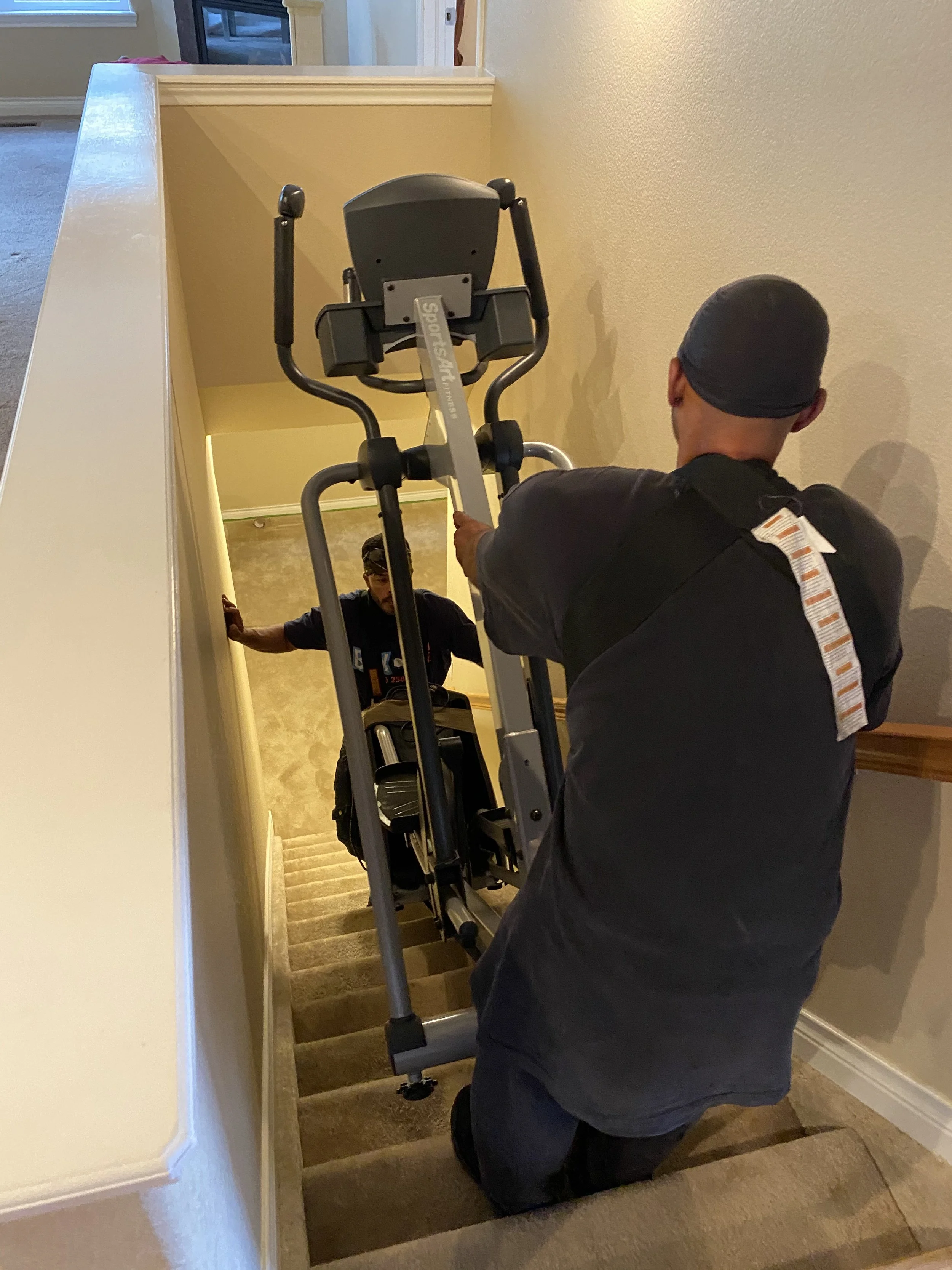 Two people moving an elliptical machine up a staircase in a house.