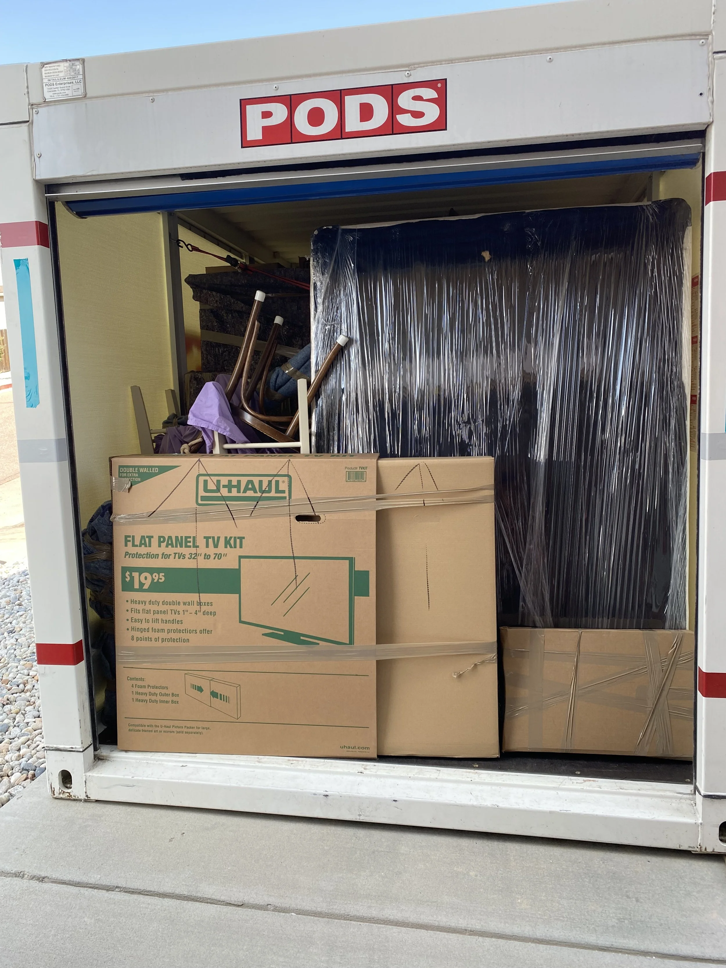 A storage PODS container filled with moving boxes, including a U-Haul flat panel TV kit and various furniture wrapped in plastic.