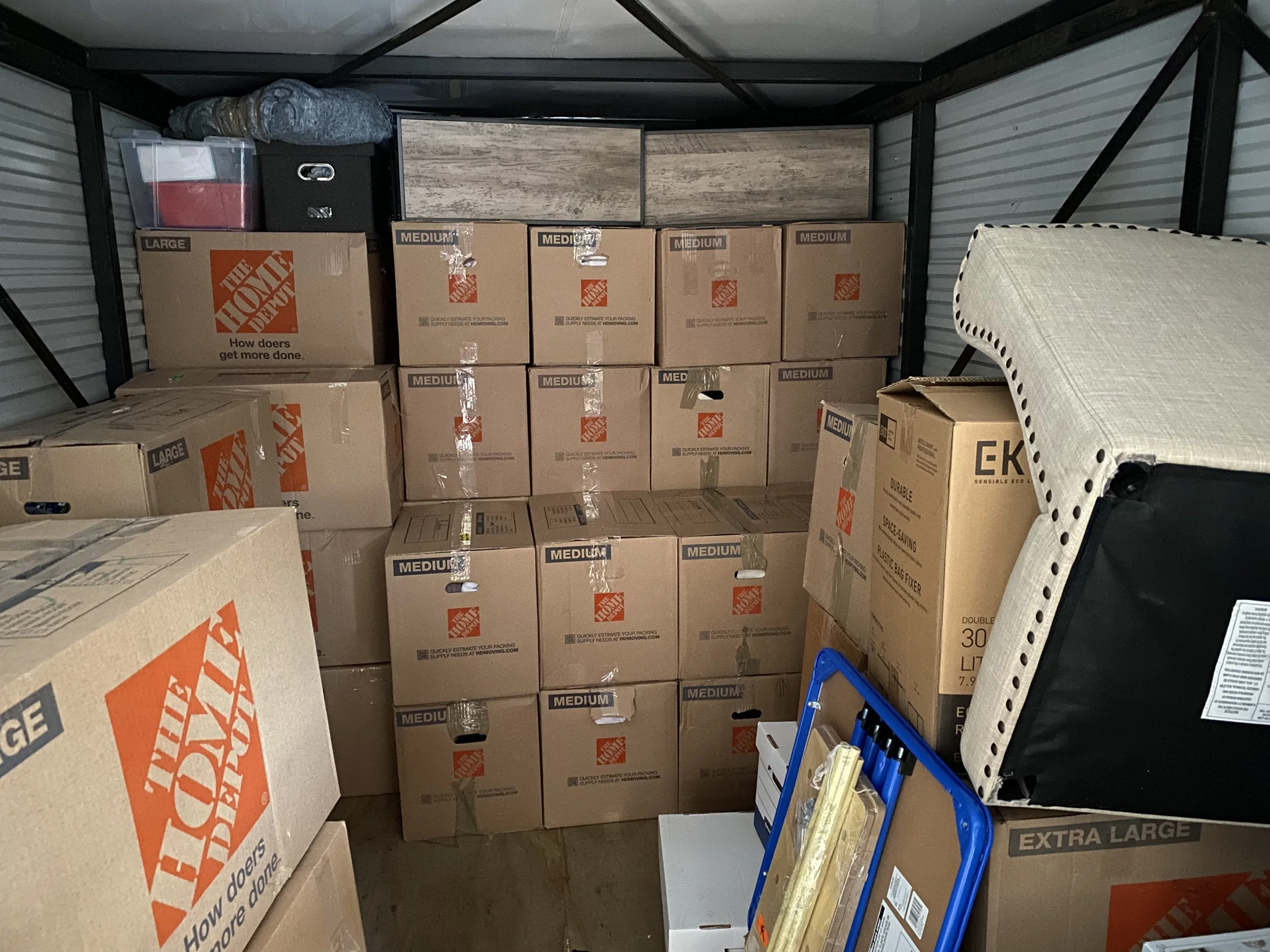Stacked moving boxes in a storage unit with blankets and furniture items.
