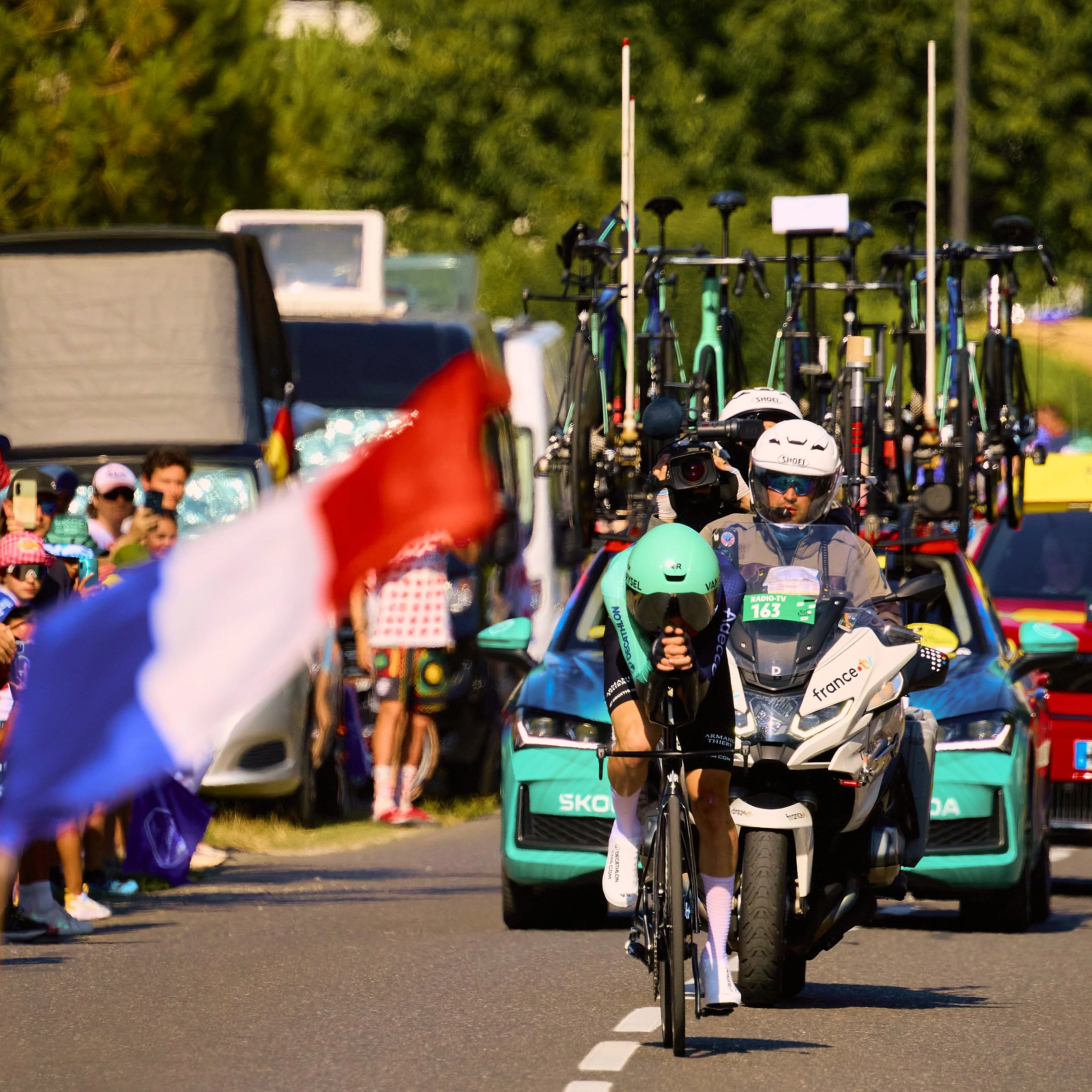Coureur suivi par la moto télévision, drapeau français au premier plan, Tour de France 2026 @Paul SEIXAS
