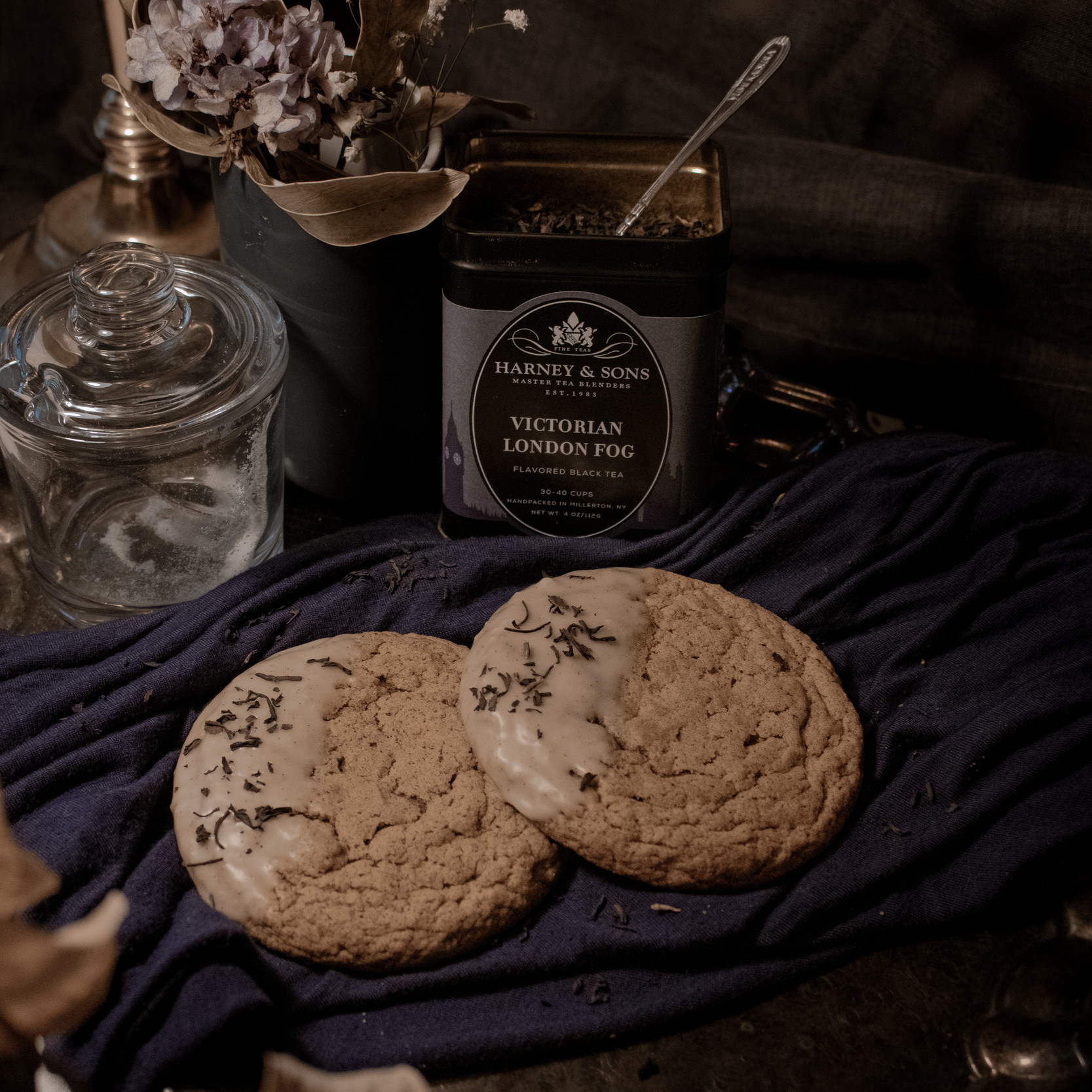 Cookies with white icing and sprinkles resting on a dark cloth, a jar of sugar, a container of black tea labeled 'Victorian London Fog,' a glass cup, and a small bouquet of dried flowers in a black vase in the background.